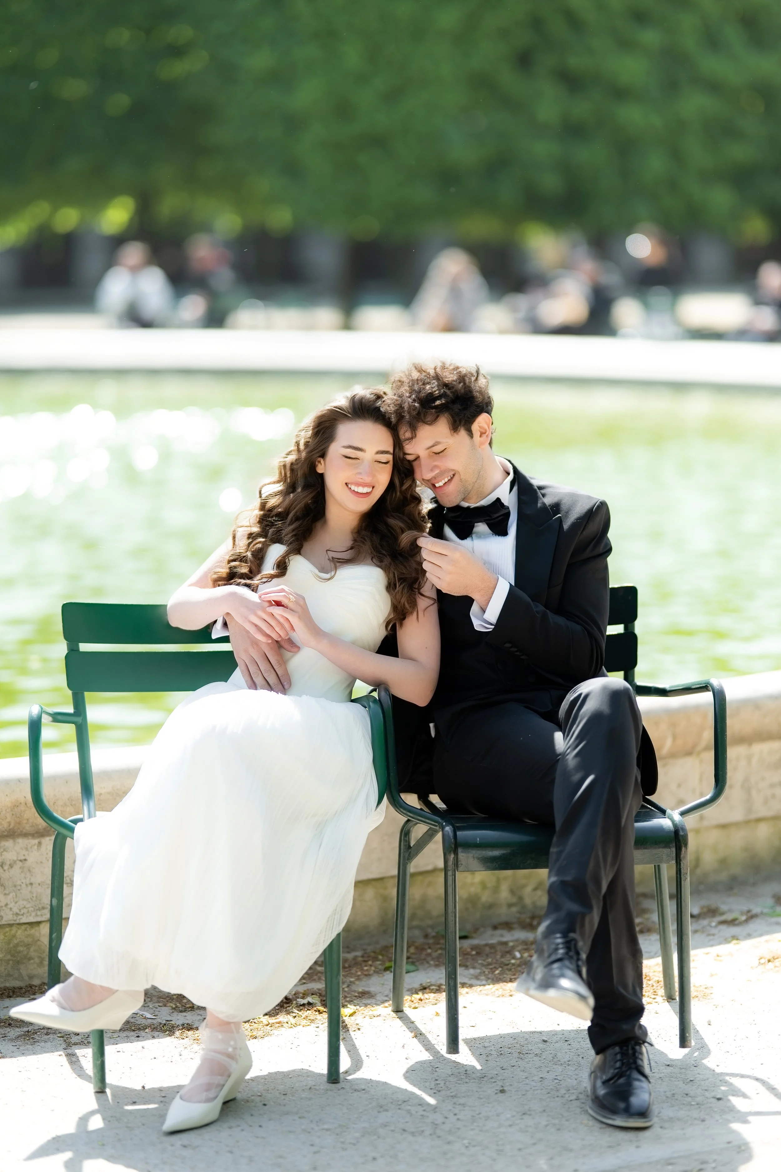 A newlywed couple sitting on a park bench, smiling and enjoying a sunny day by a lake, with a lush green background and other people in the distance.