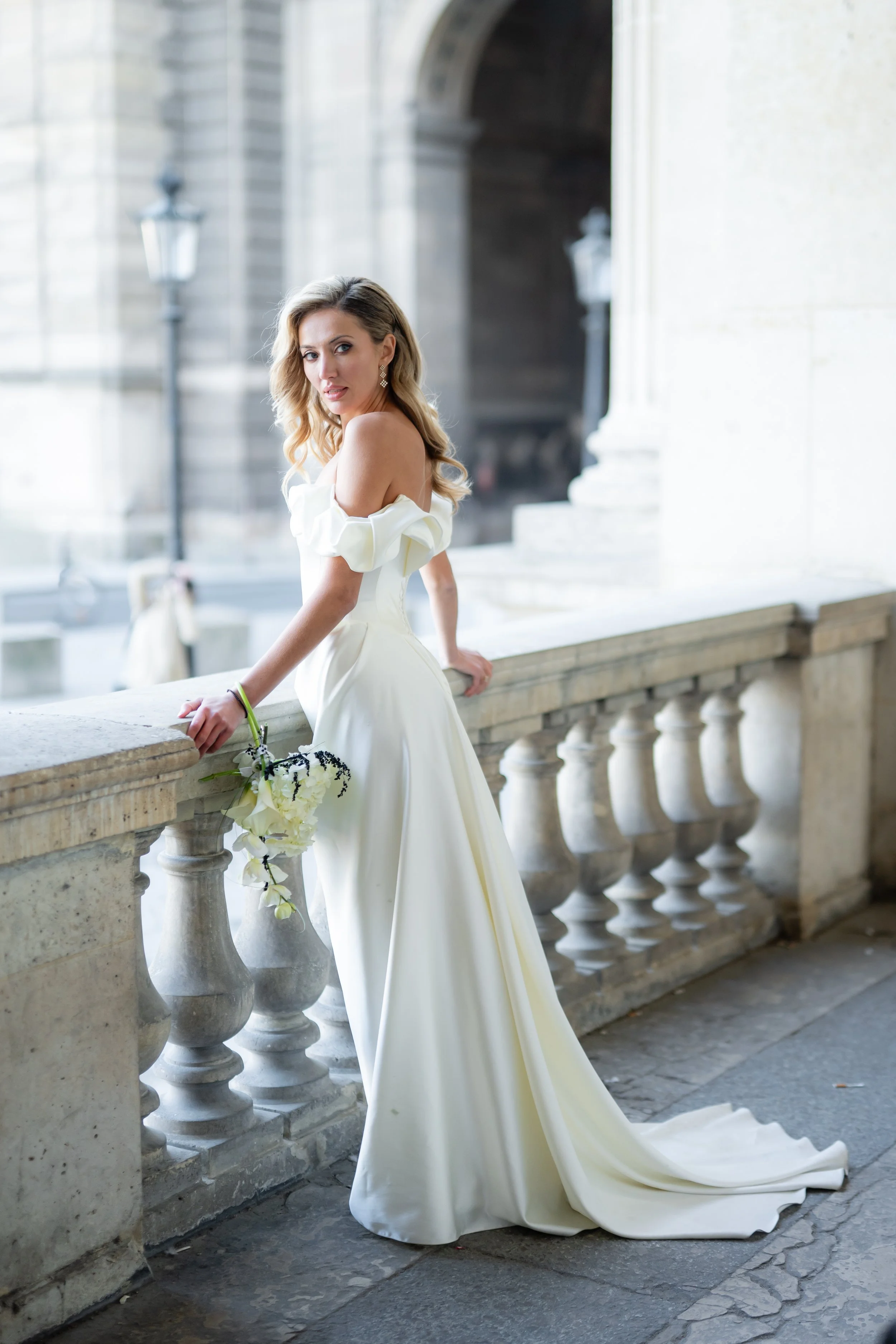 A woman in a white wedding dress, with loose blonde hair, and statement earrings, leaning against a stone balustrade on a historic building's balcony, holding a floral bouquet.