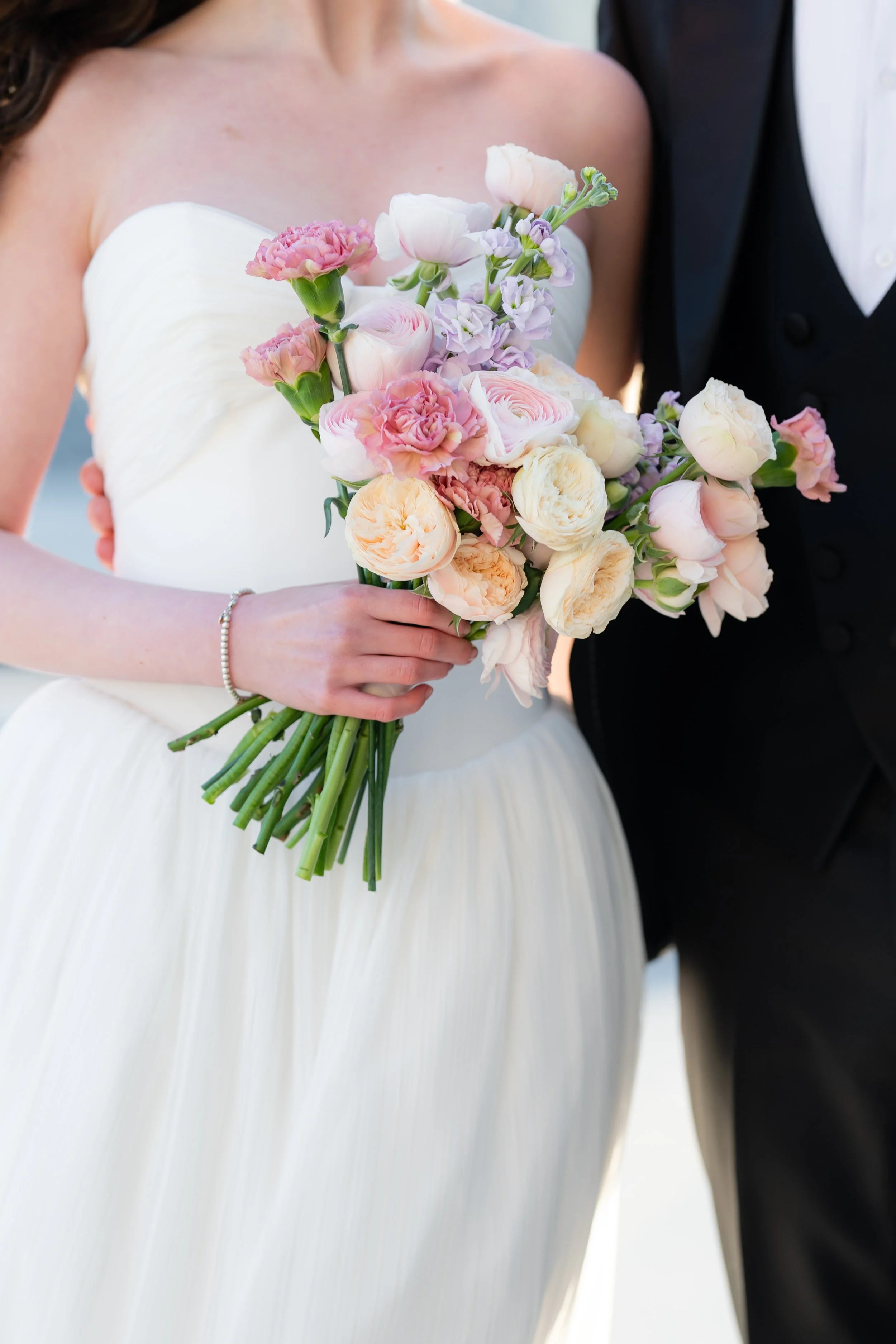 A bride wearing a white strapless wedding dress holding a bouquet of pink, white, and light purple flowers, with a groom standing beside her in a black suit.
