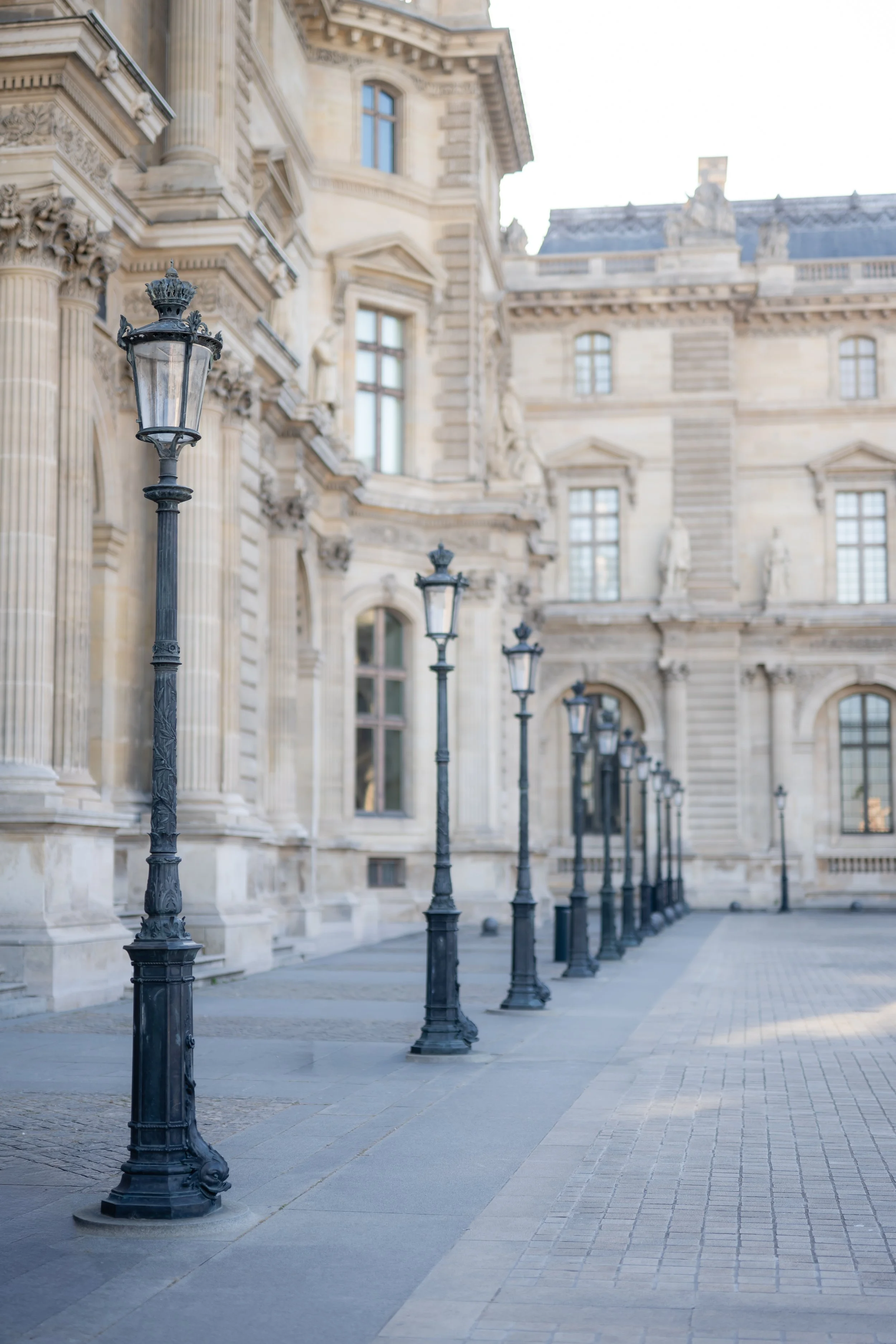 Row of vintage black street lamps lining a sidewalk in front of an ornate historic building with large windows and detailed architectural decorations.