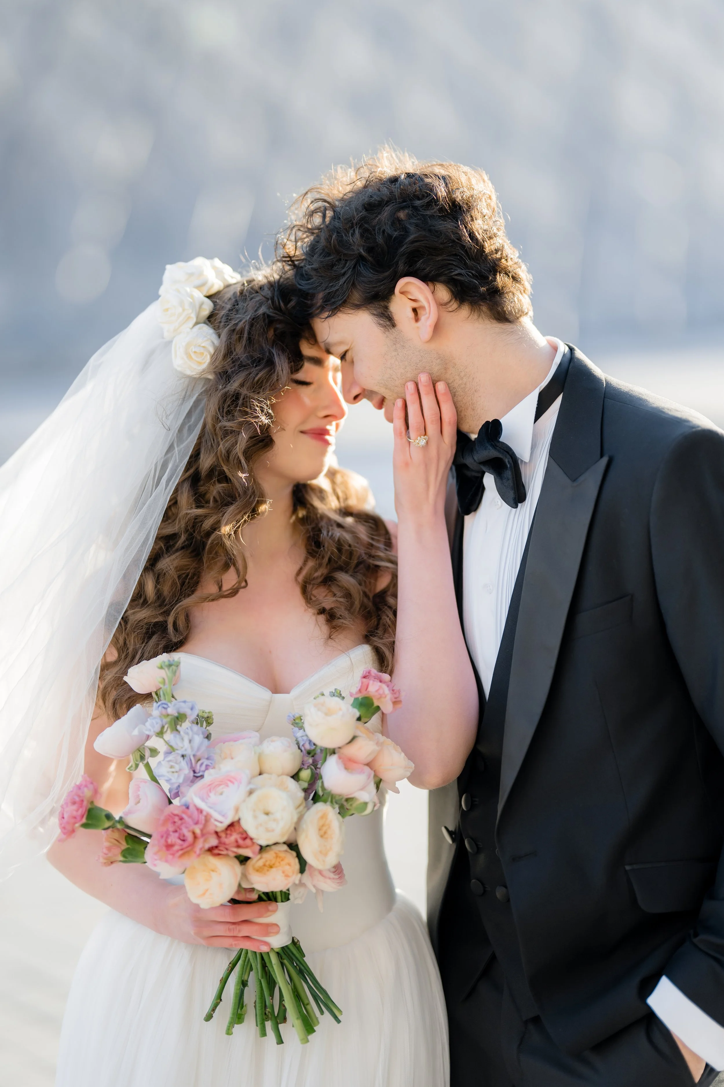 A bride and groom share a tender moment, with their foreheads touching and eyes closed. The bride is holding a colorful bouquet of flowers and is wearing a white wedding dress and veil adorned with flowers. The groom is dressed in a black tuxedo with