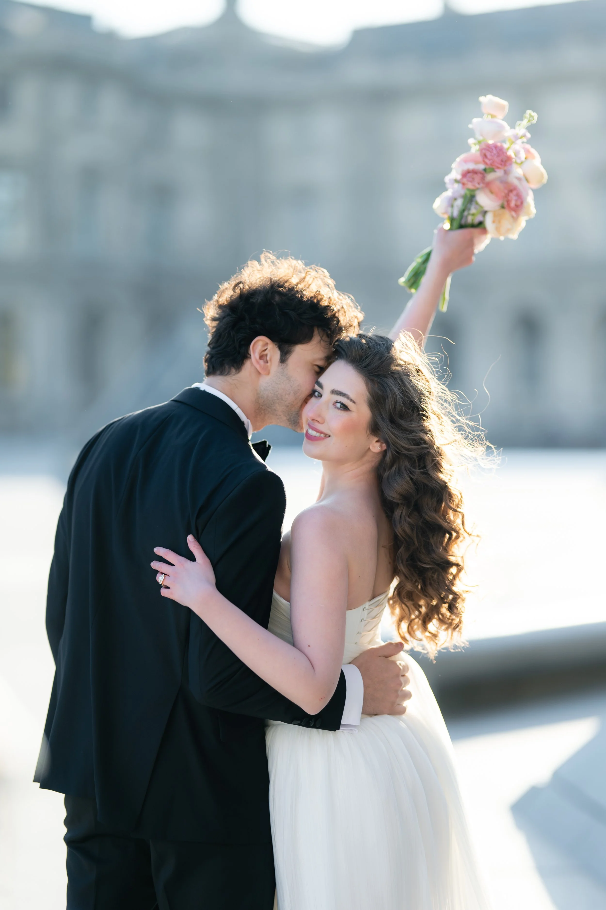 A newlywed couple embraces outdoors near the Louvre, with the bride holding a bouquet of pink and white flowers and the groom kissing her forehead, during sunset or sunrise.