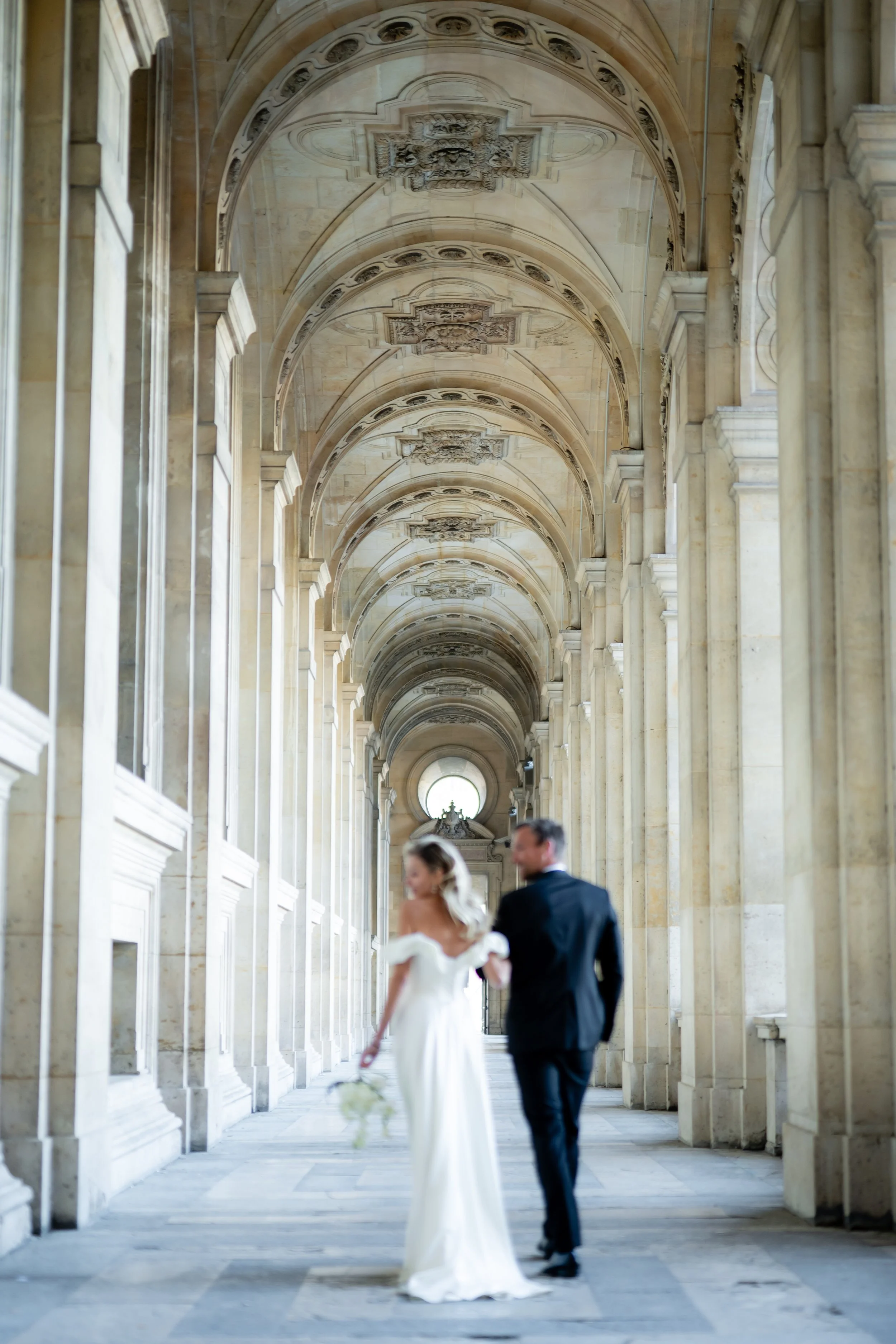 A bride and groom walking through an elegant, arched, colonnaded hallway with detailed ceiling architecture, in a historic or classical building, when their faces are blurred.