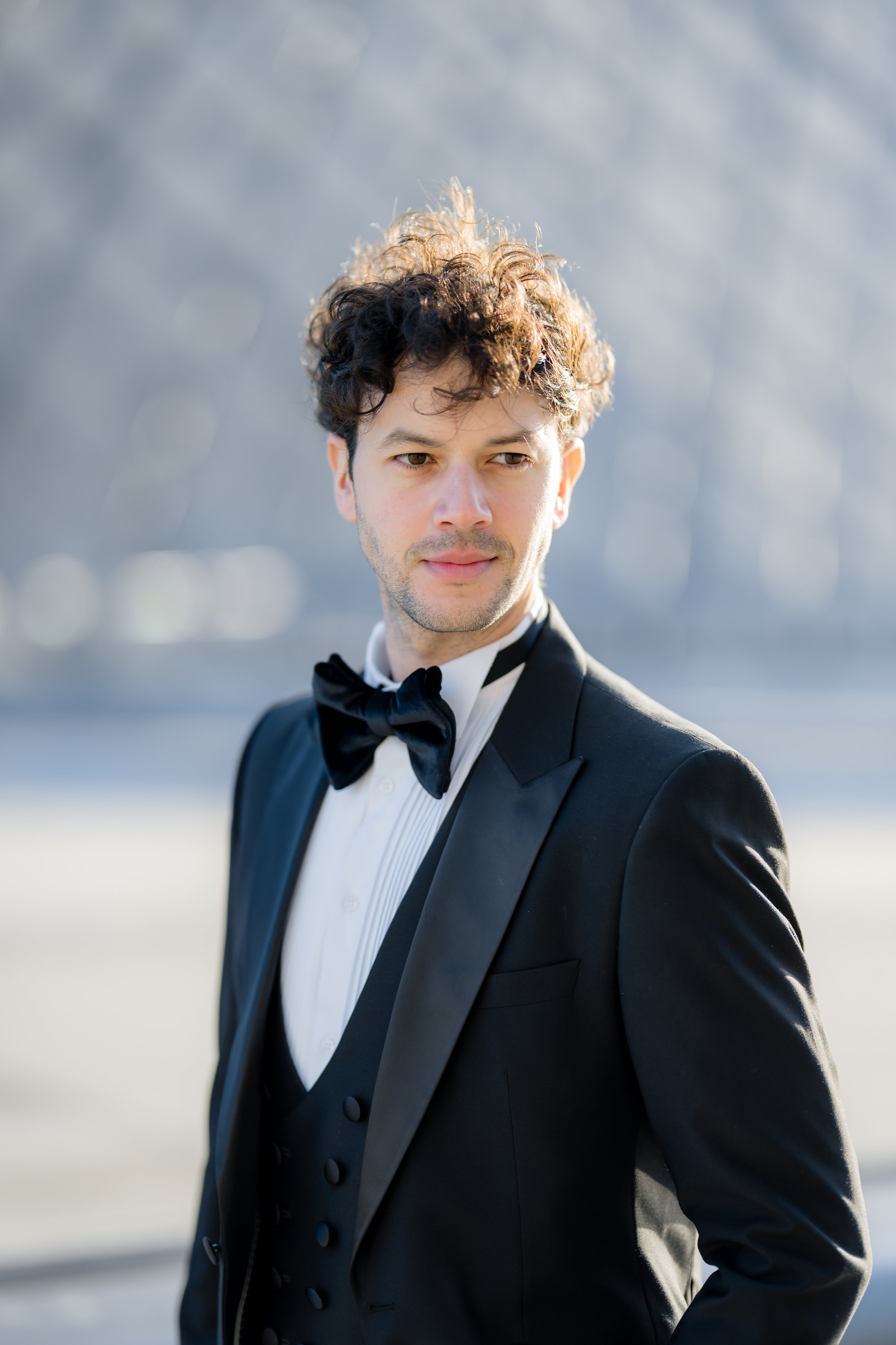 A man in a black tuxedo with a bow tie standing outdoors with a blurred background.