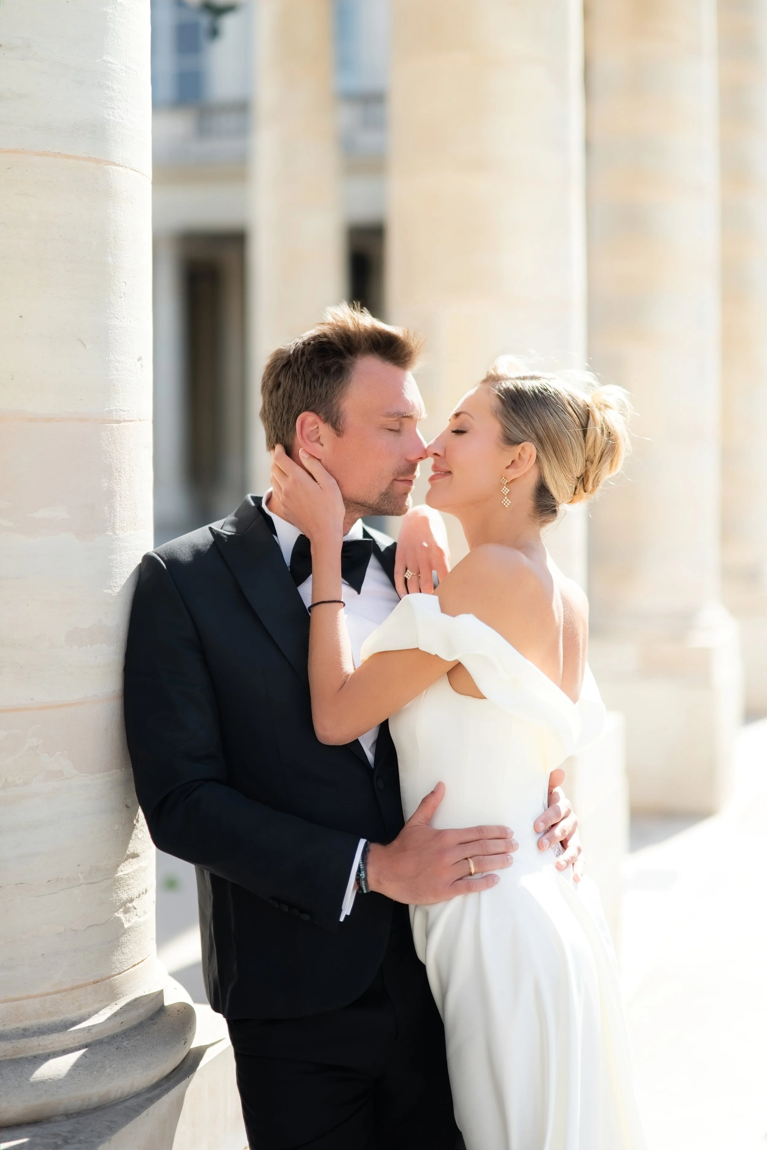 A man and woman in wedding attire sharing an intimate moment outdoors near stone pillars, with the man dressed in a tuxedo and the woman in a white wedding dress.