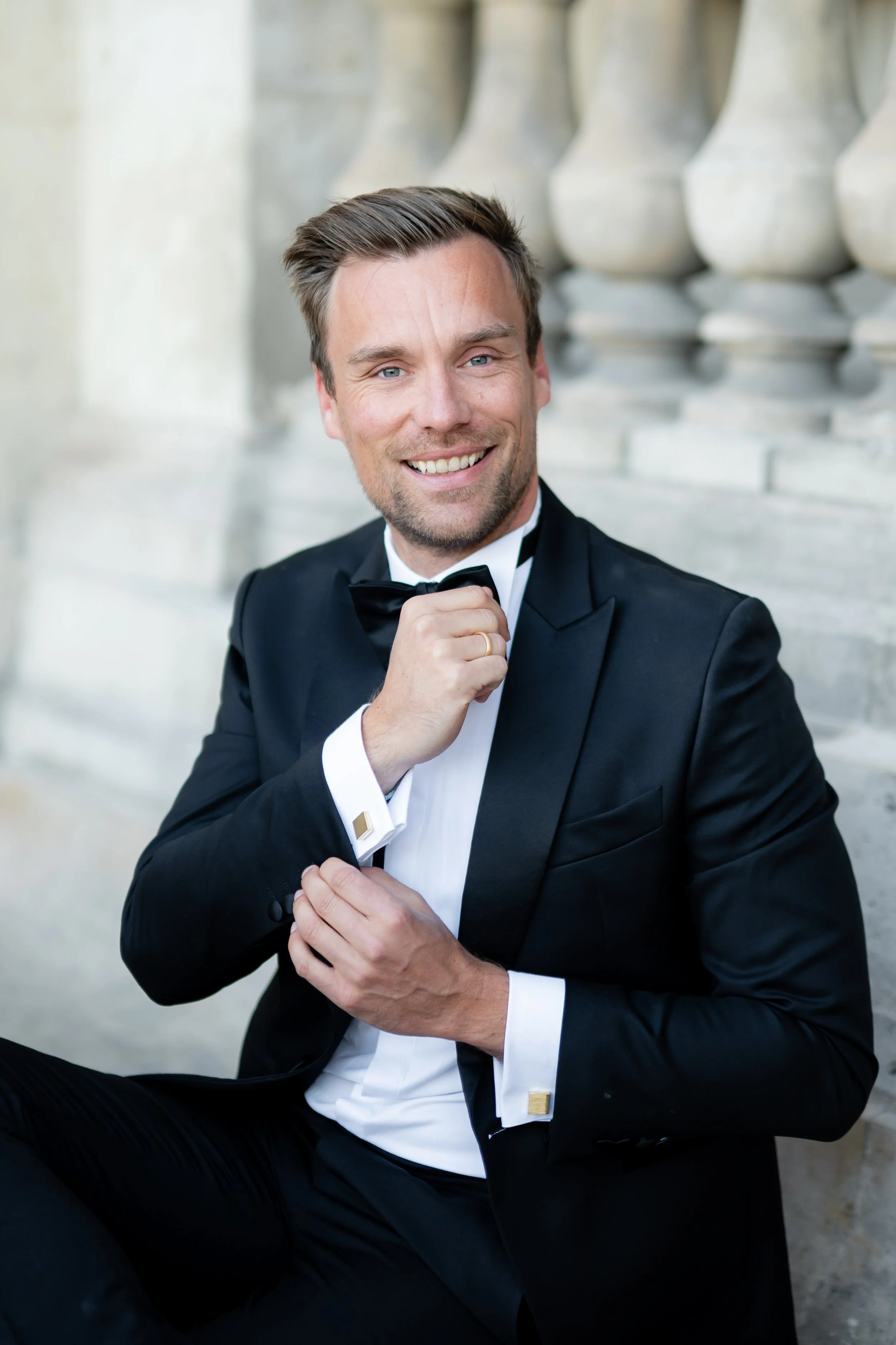 A man in a black tuxedo with a bow tie, smiling, and adjusting his collar, seated outdoors against a stone wall with classical columns.