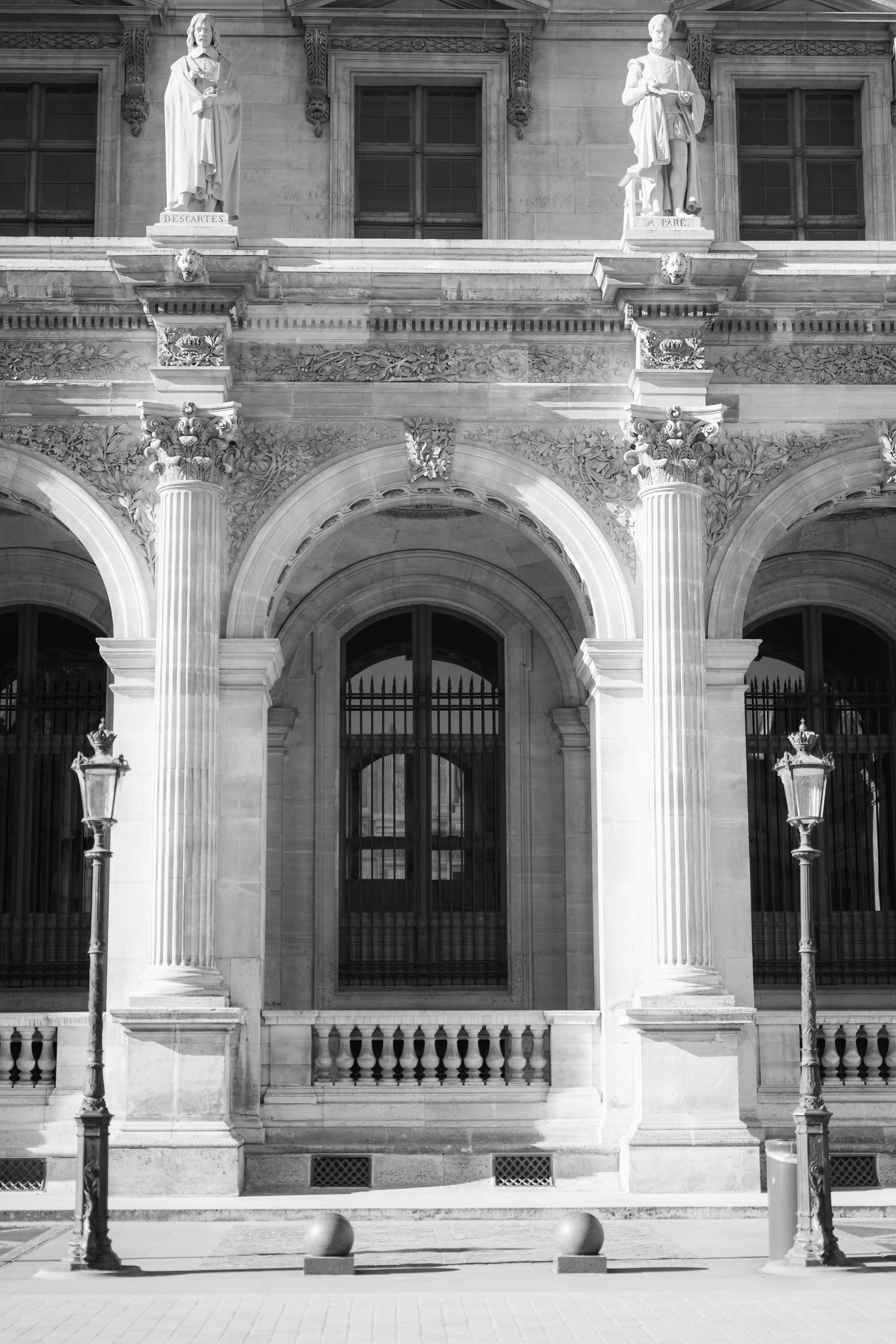 Black and white photograph of the facade of a historic building with two statues on plinths above a large arched entrance, iron gates, and two street lamps in front of the building.
