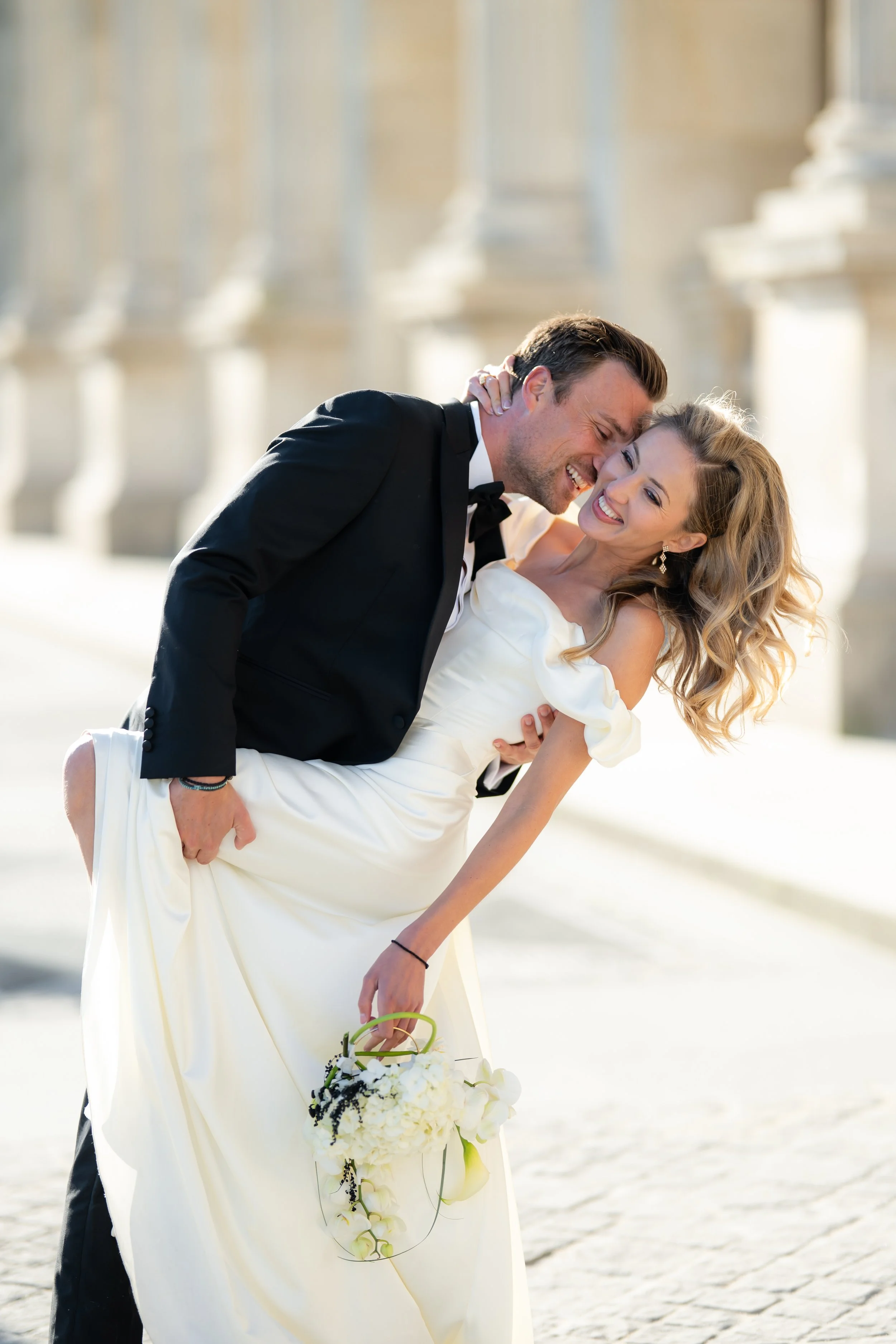 A couple dressed in formal wedding attire, with the man in a black tuxedo and the woman in a white wedding dress, smiling and sharing a joyful moment outdoors on a sunny day.