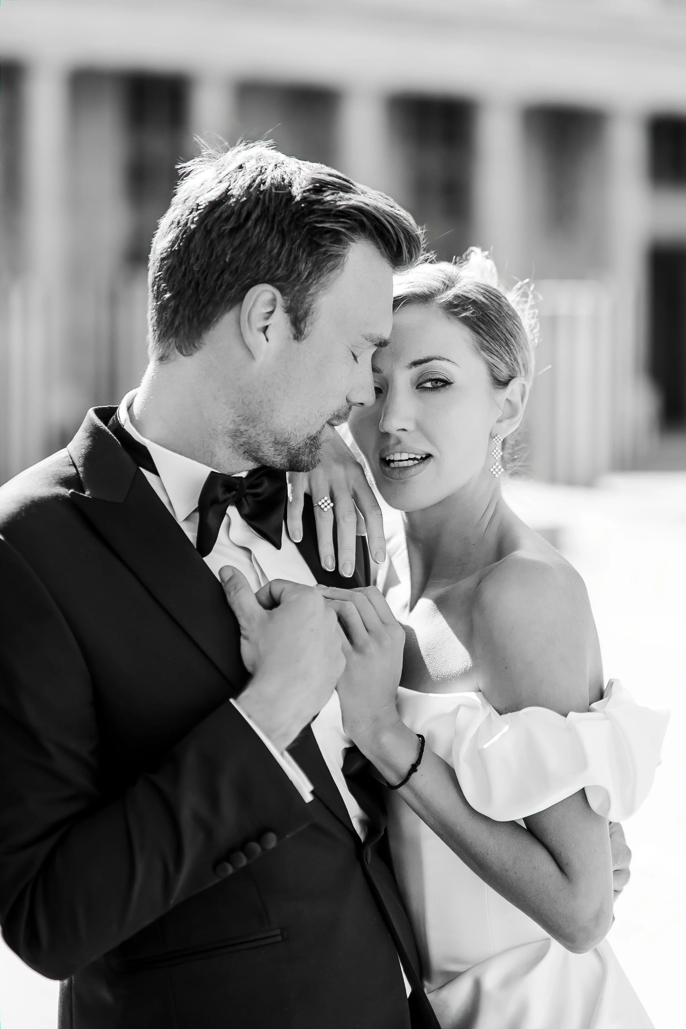 A black-and-white photo of a man in a tuxedo and a woman in a wedding dress, holding hands and touching foreheads, with a blurred building background.