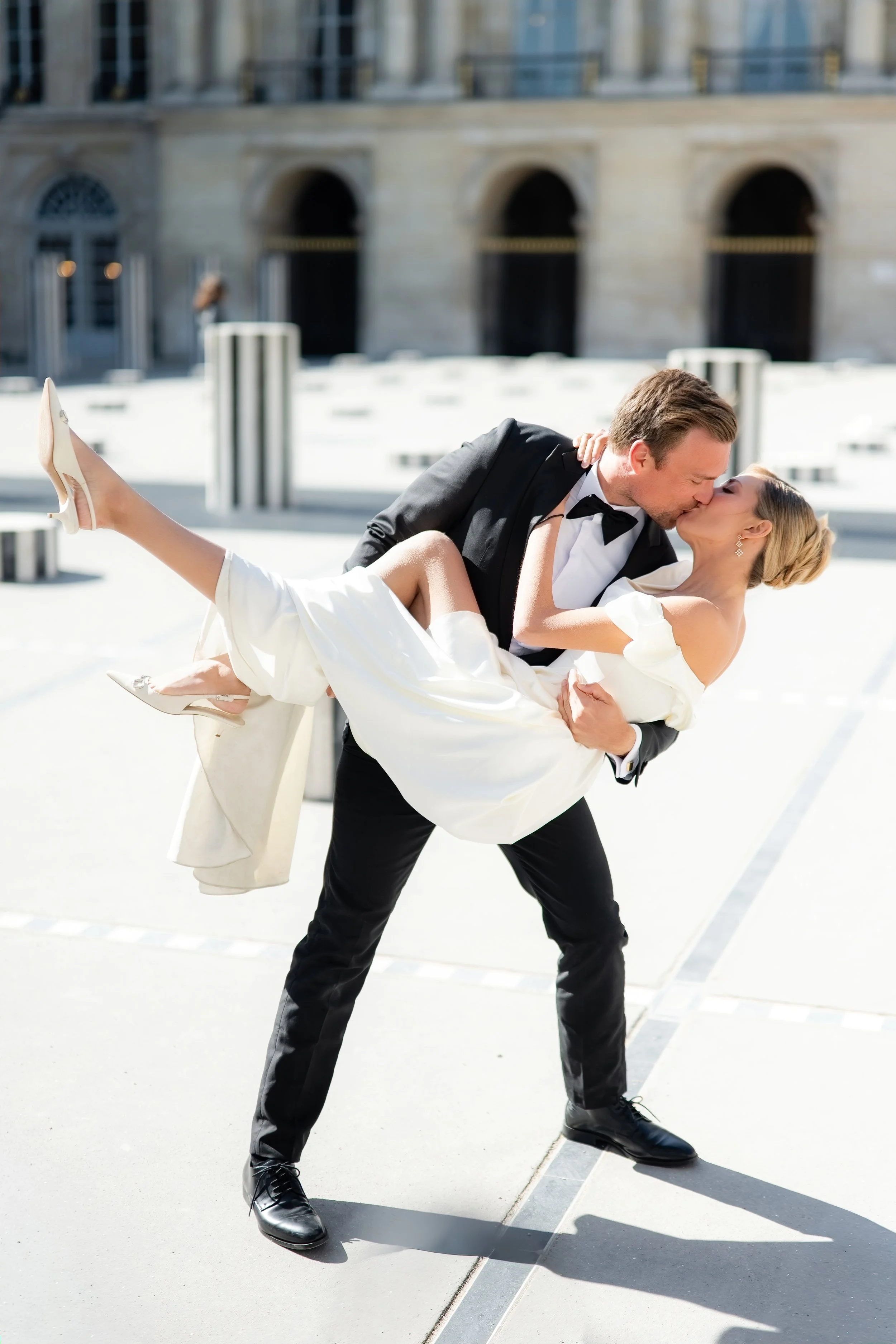 A man in a tuxedo is holding up a woman in a white dress and high heels, dancing outdoors on a city street with a large building in the background.