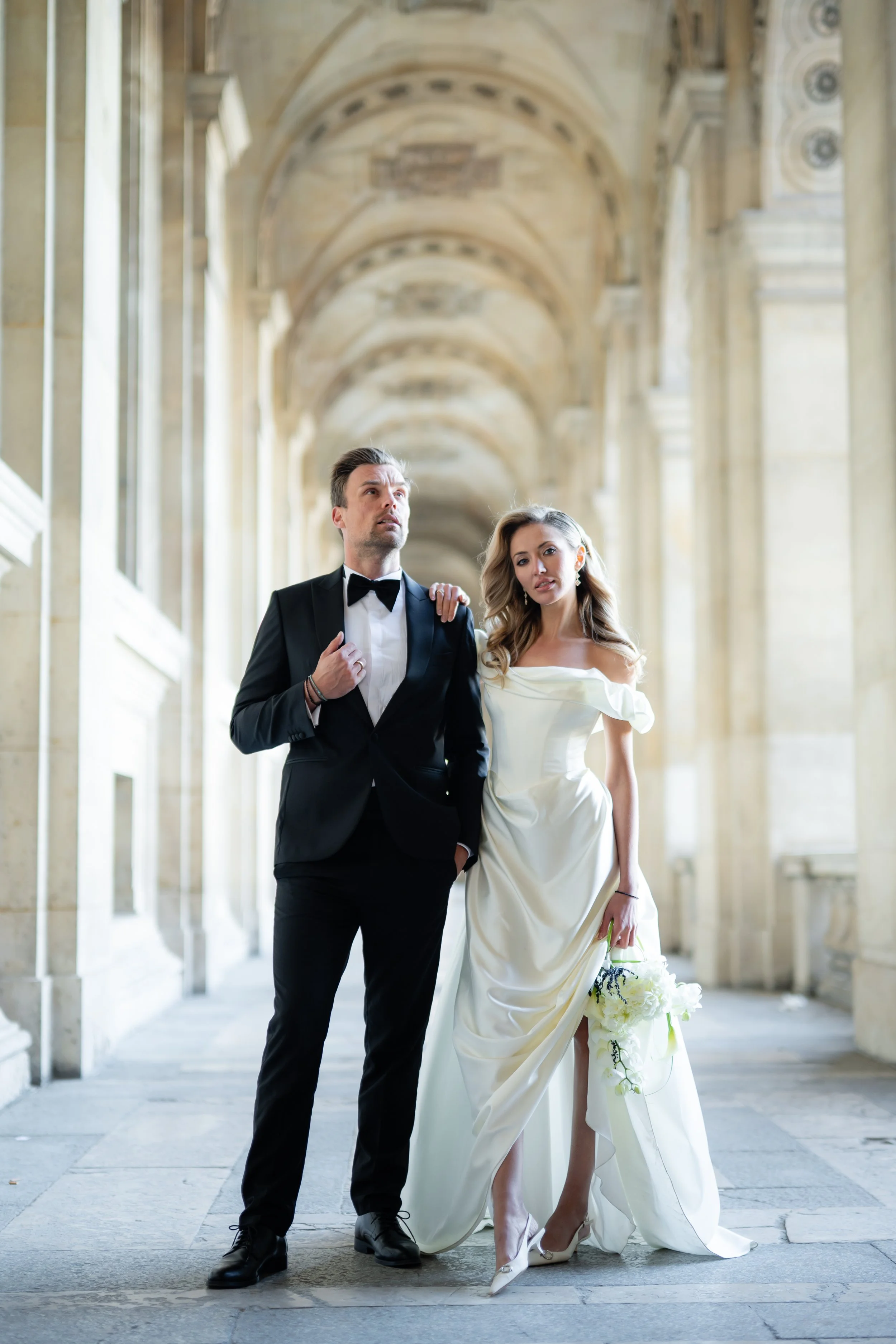 A man in a tuxedo and a woman in a white wedding dress standing in an elegant, classical architectural corridor with high arches and columns.