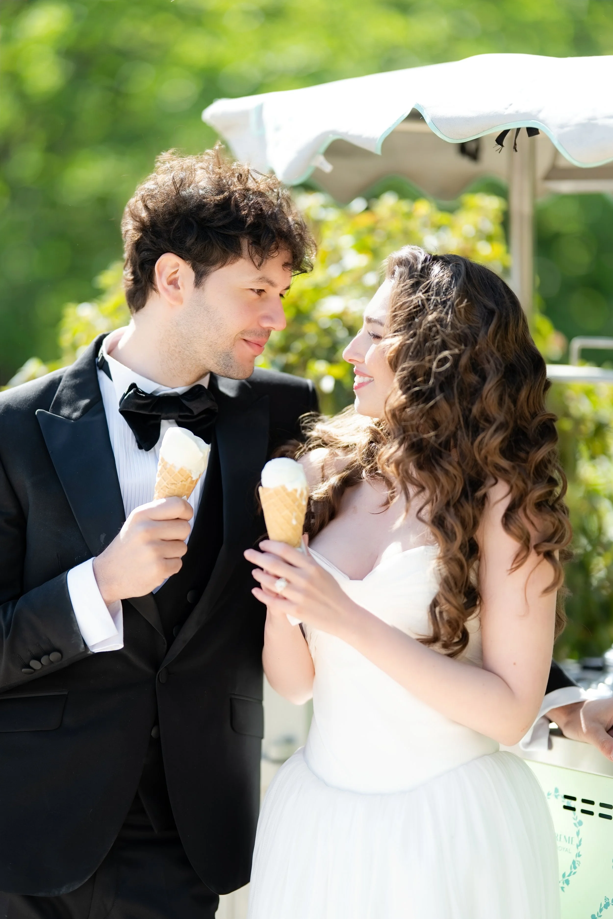A newlywed couple in formal wedding attire, with the groom in a tuxedo and the bride in a white wedding dress, enjoying ice cream cones outdoors under a sunny sky.