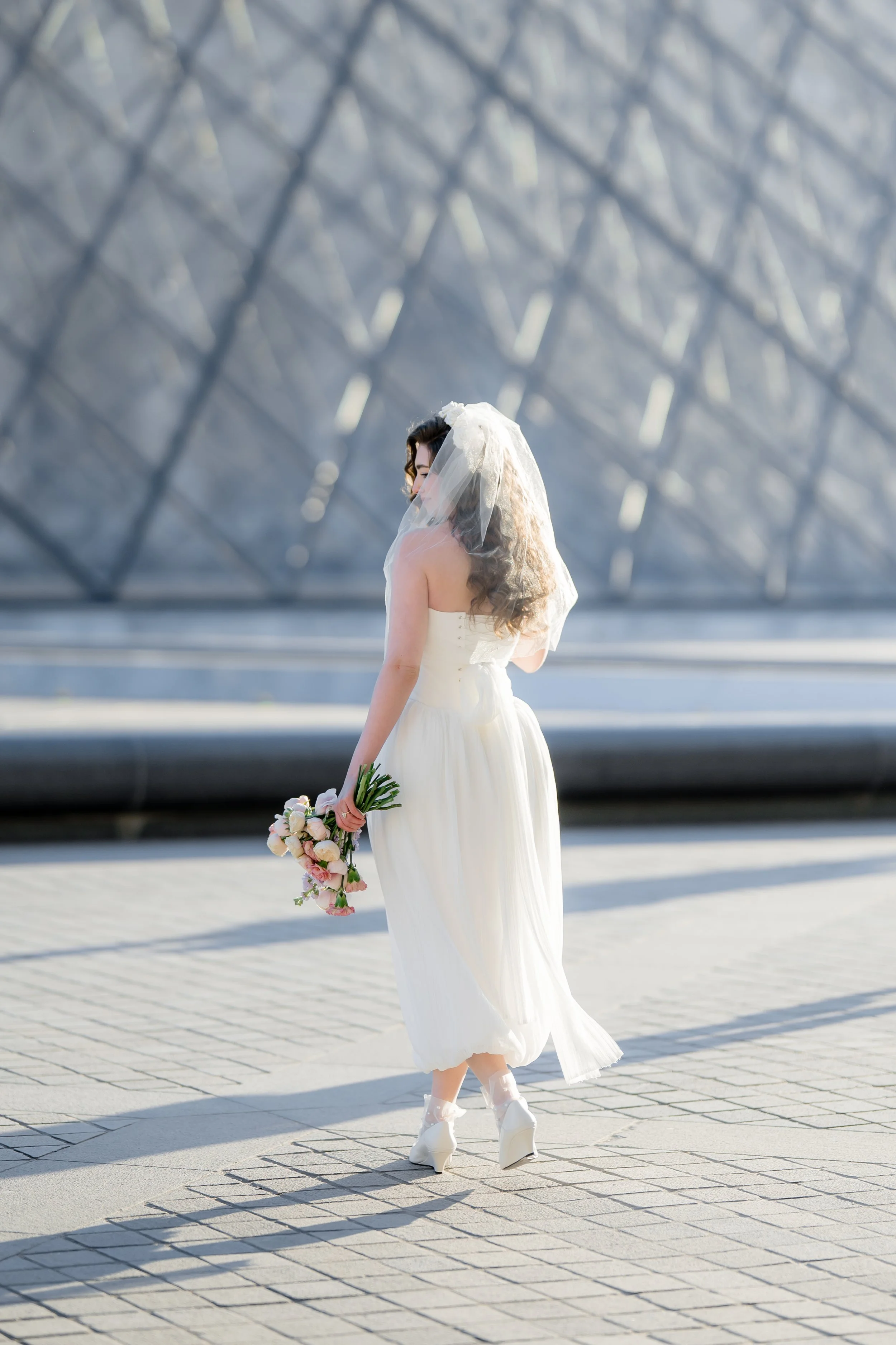 A bride in a white dress and veil holding a flower bouquet standing outdoors on a paved area in front of the louvre