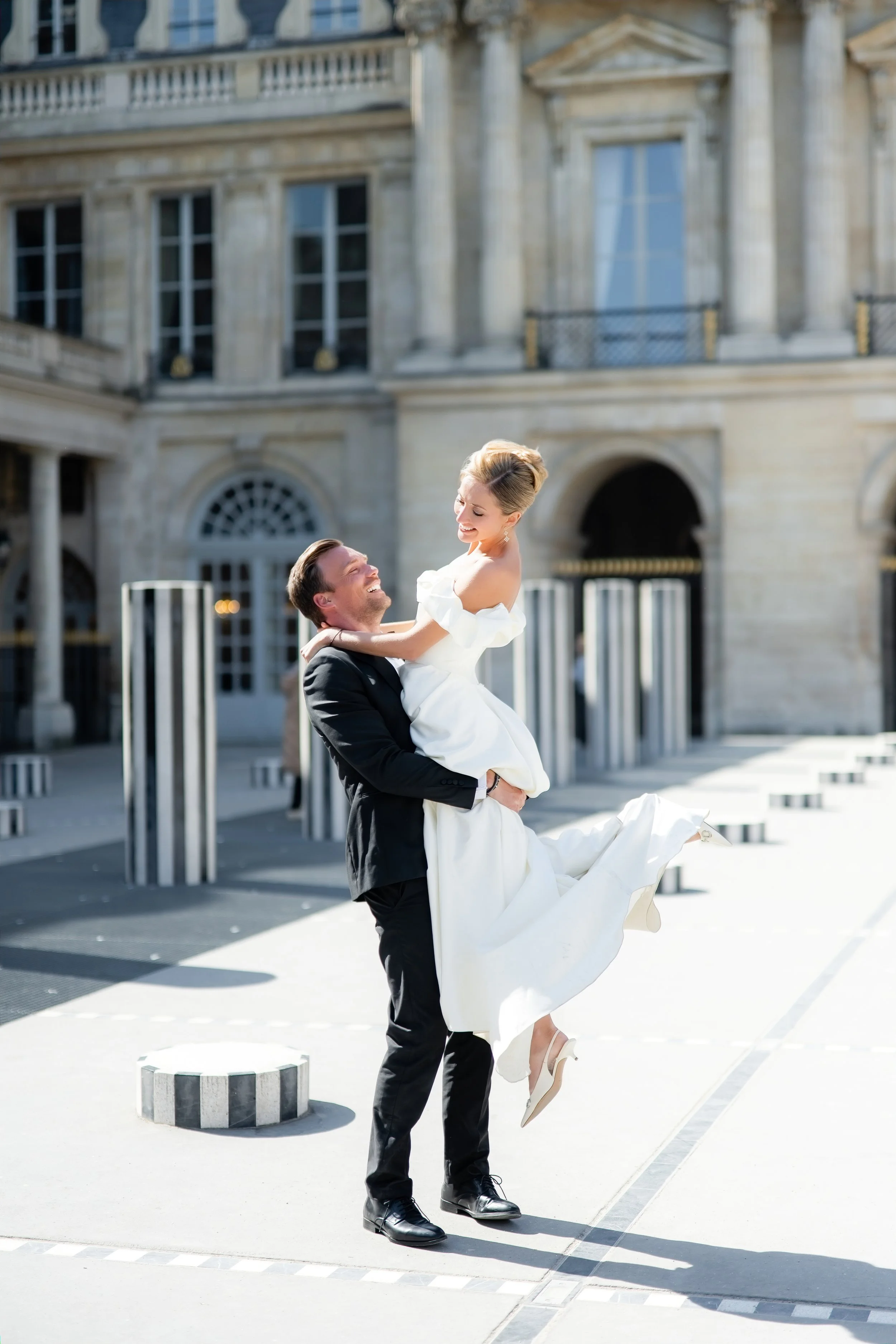 A man in a black suit lifting a woman in a white dress in an outdoor urban setting with a grand historic building in the background.