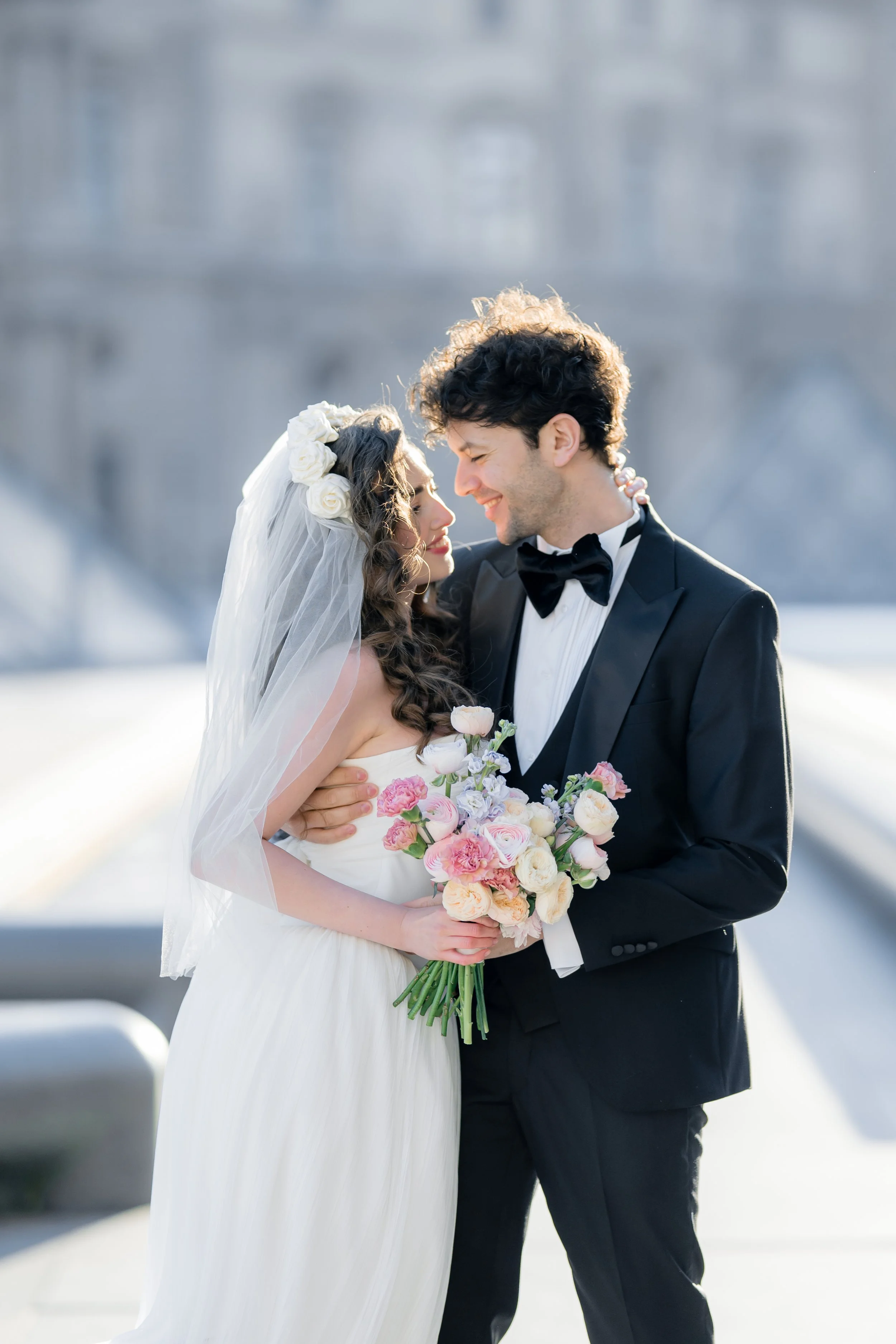 A bride and groom in wedding attire smiling and holding each other outdoors on a sunny day, with the bride holding a bouquet of pink and white flowers.