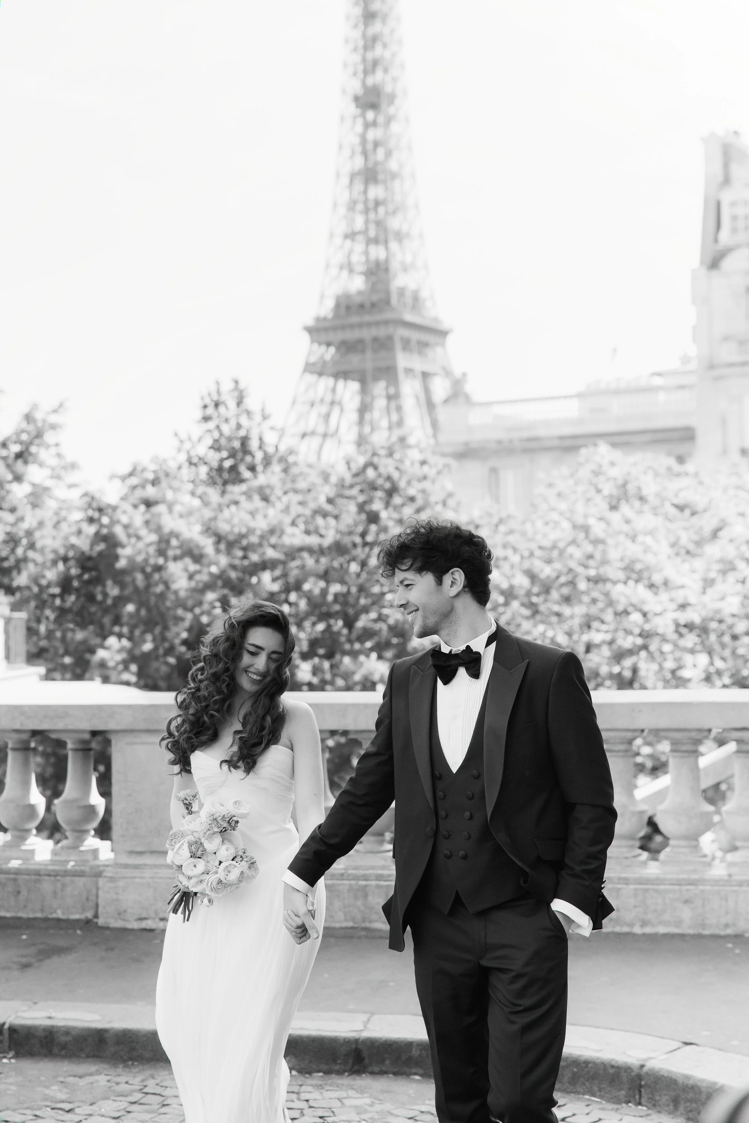 A bride and groom walking hand in hand outdoors in front of the Eiffel Tower, smiling and looking at each other.