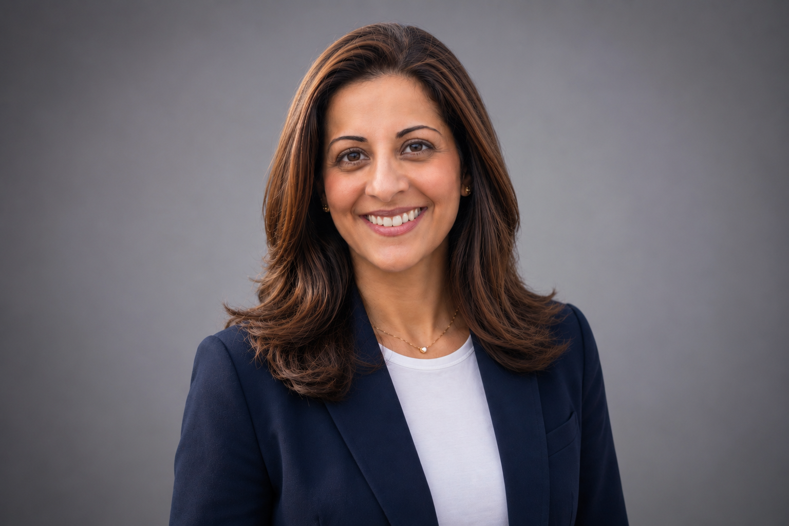 A woman with medium-length brown hair, smiling, wearing a navy blazer and a white top, standing against a plain gray background.