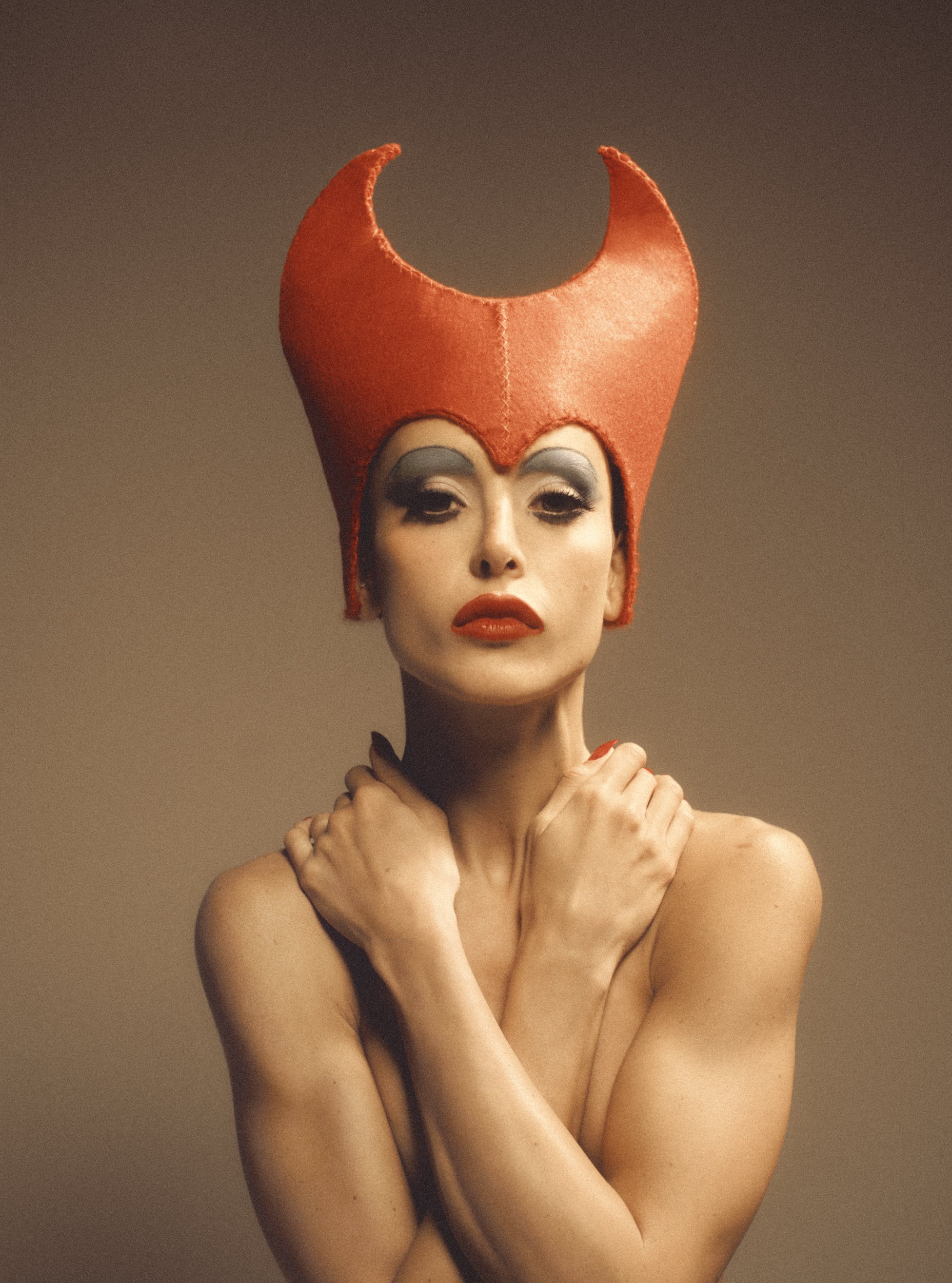 A woman with dramatic makeup and a red, horned hat, holding her arms crossed over her chest.