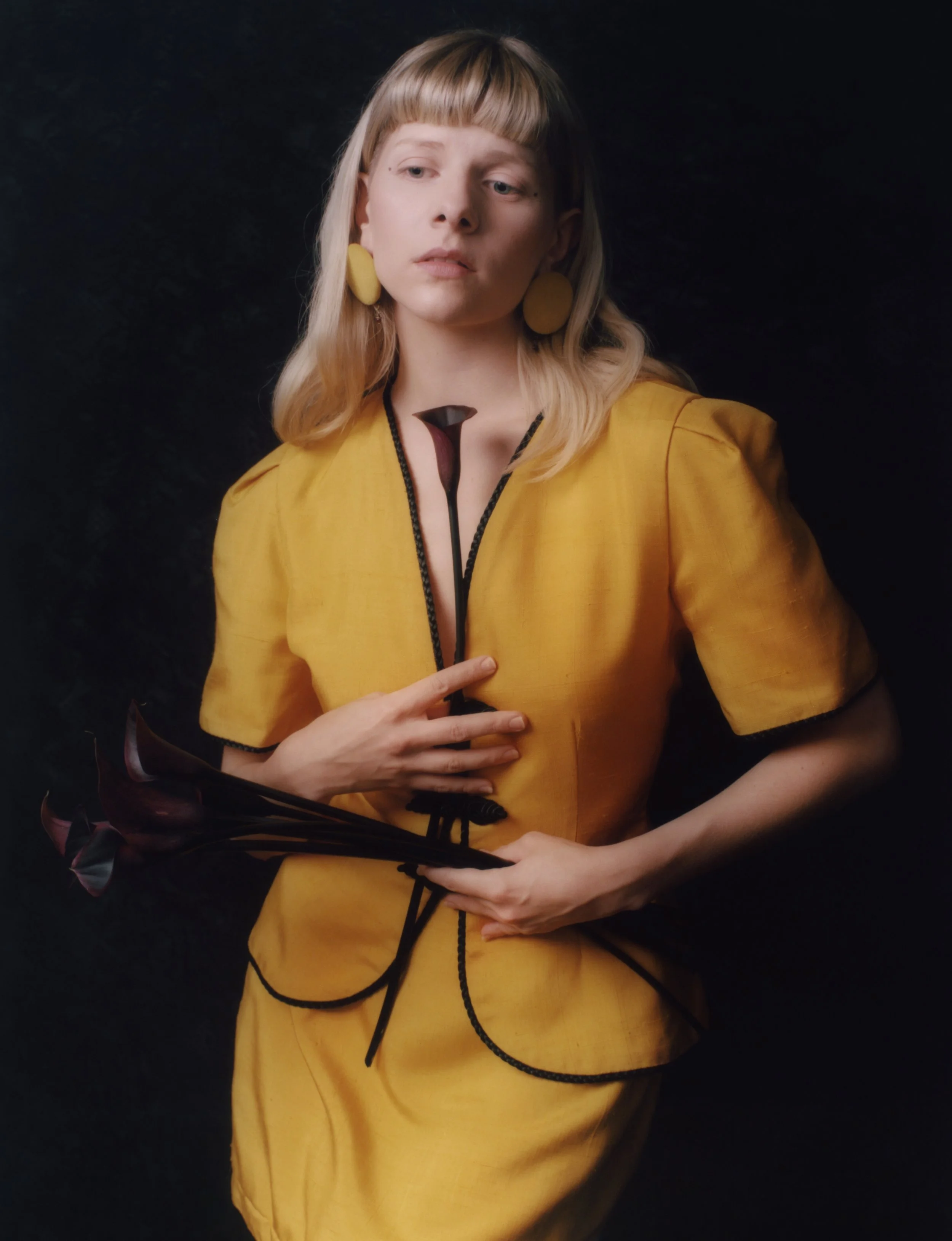 A young woman with blonde hair and bangs, wearing a yellow dress with black accents, yellow earrings, and holding a black and pink flower against a dark background.