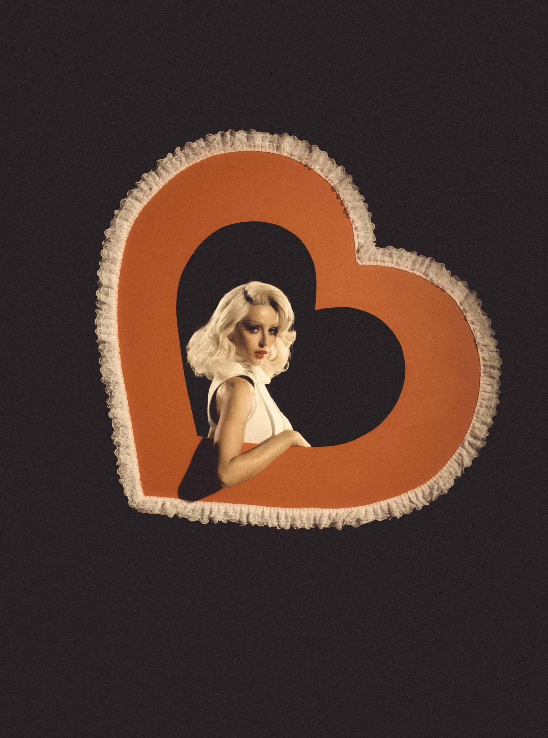 A woman with platinum blonde hair styled in vintage waves, wearing a white dress, posing in front of a large orange number 3 with a black circle inside it, on a black background.