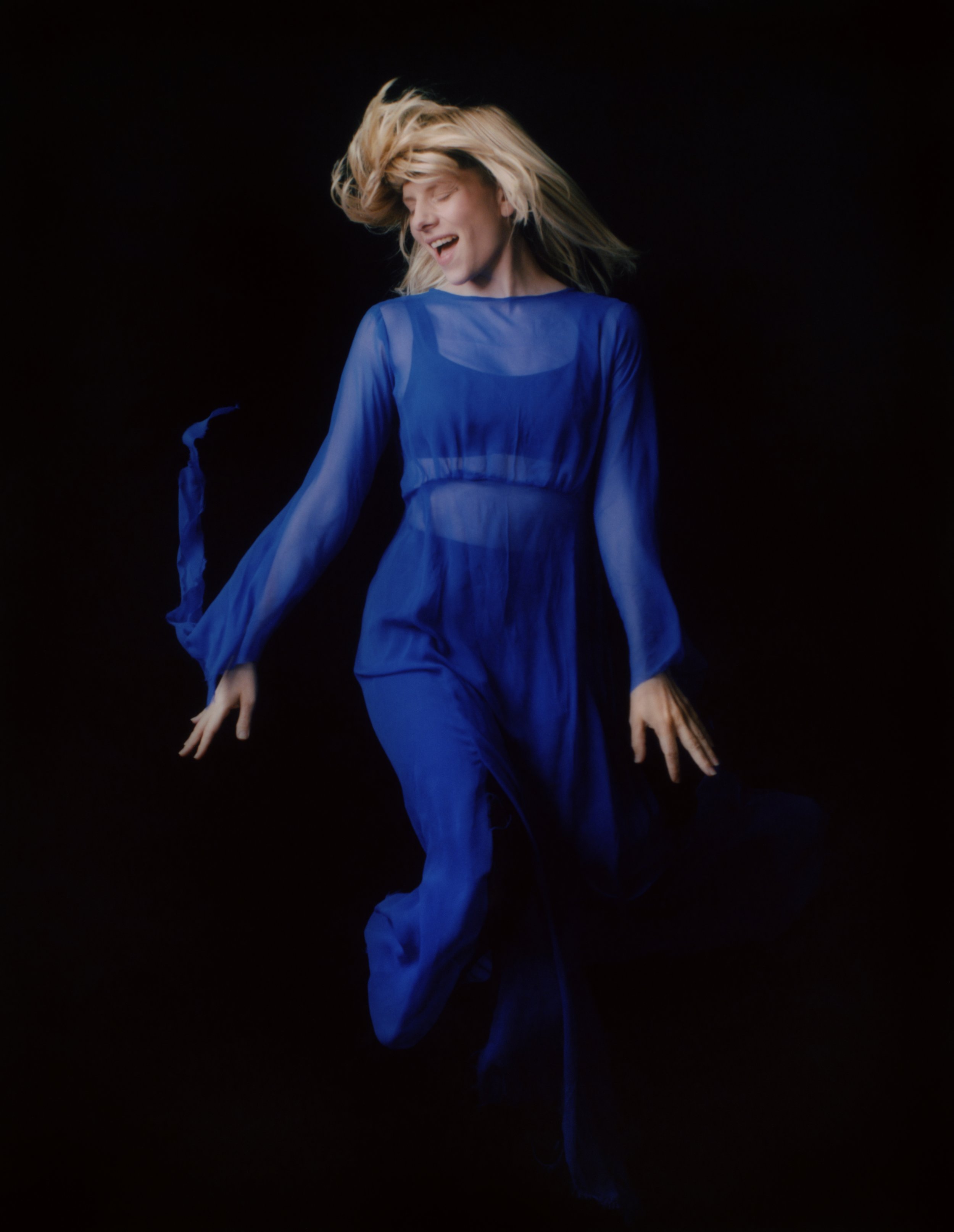 A woman with blonde hair dancing in a flowing, sheer blue dress against a black background, expression joyful.
