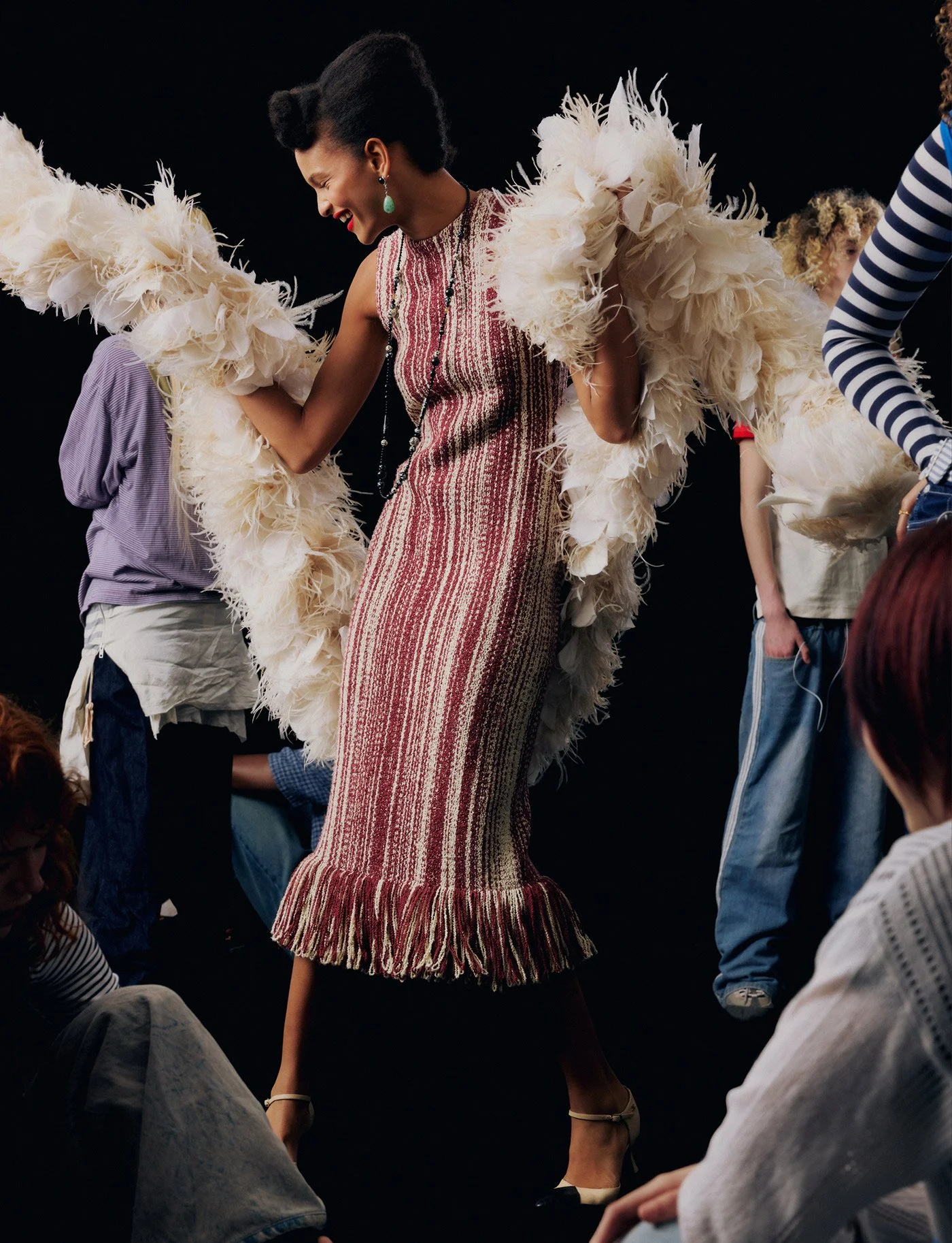 A woman in a striped dress and feathered jacket walking on a runway, surrounded by people sitting on the floor and some standing, with a black background.