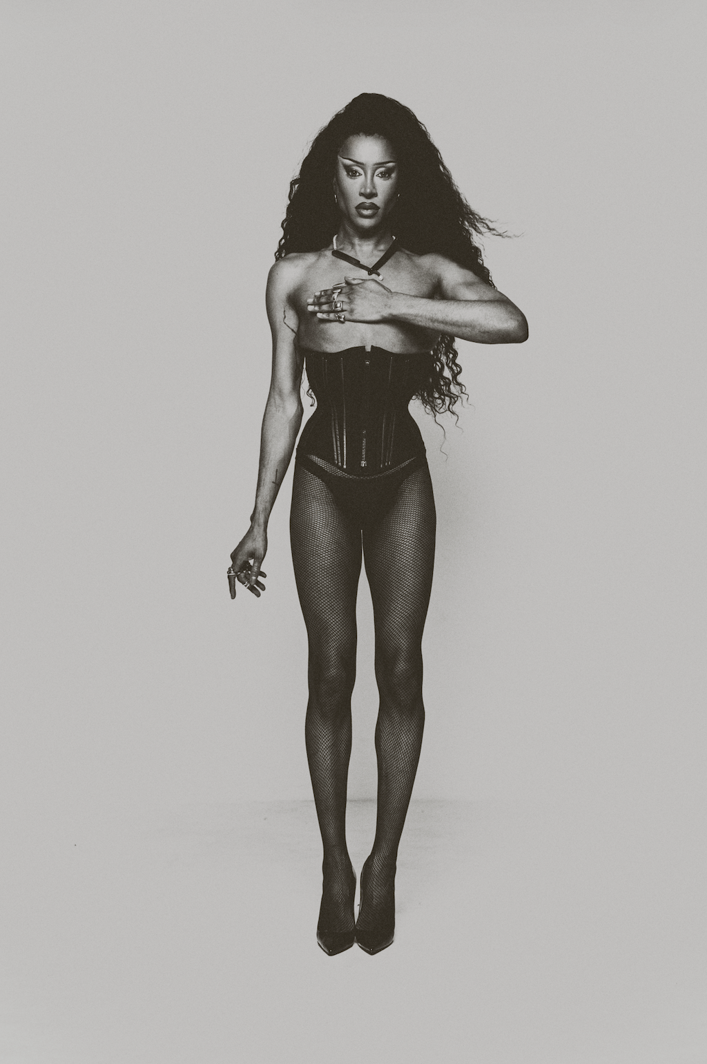 A woman with long curly hair wearing fishnet tights, high heels, a waist corset, and jewelry, standing against a plain background, holding a cigarette, and making a hand gesture with her fingers.