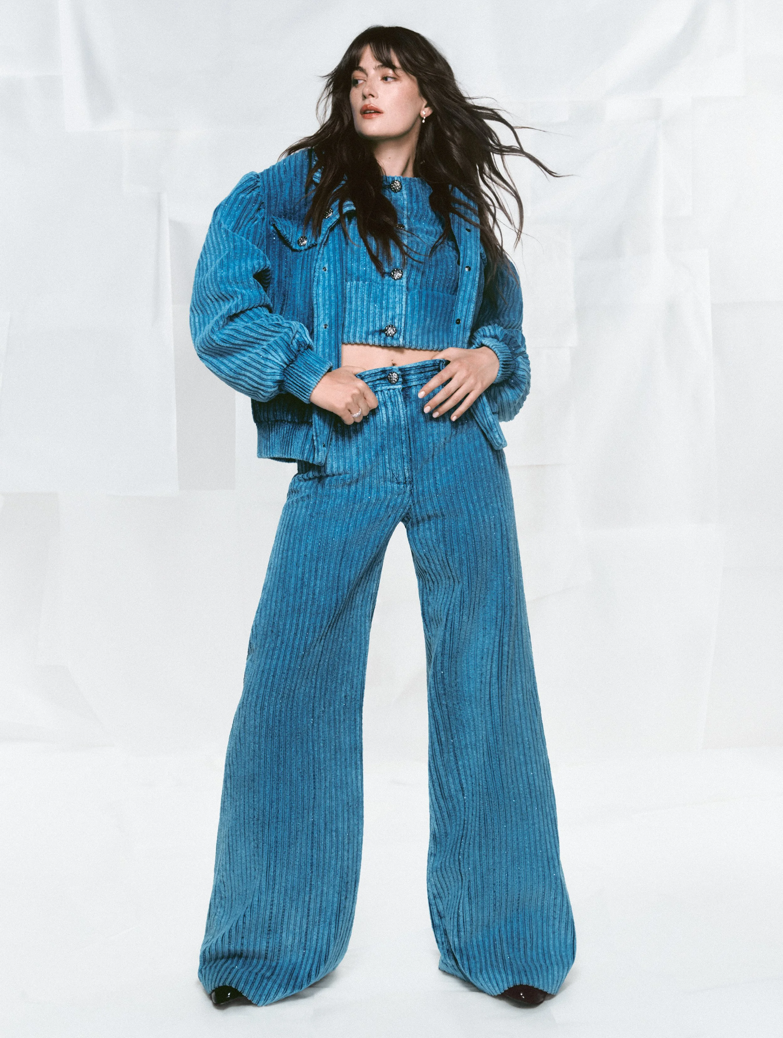 Woman with long dark hair wearing a matching blue corduroy jacket and wide-leg pants, standing against a light background.