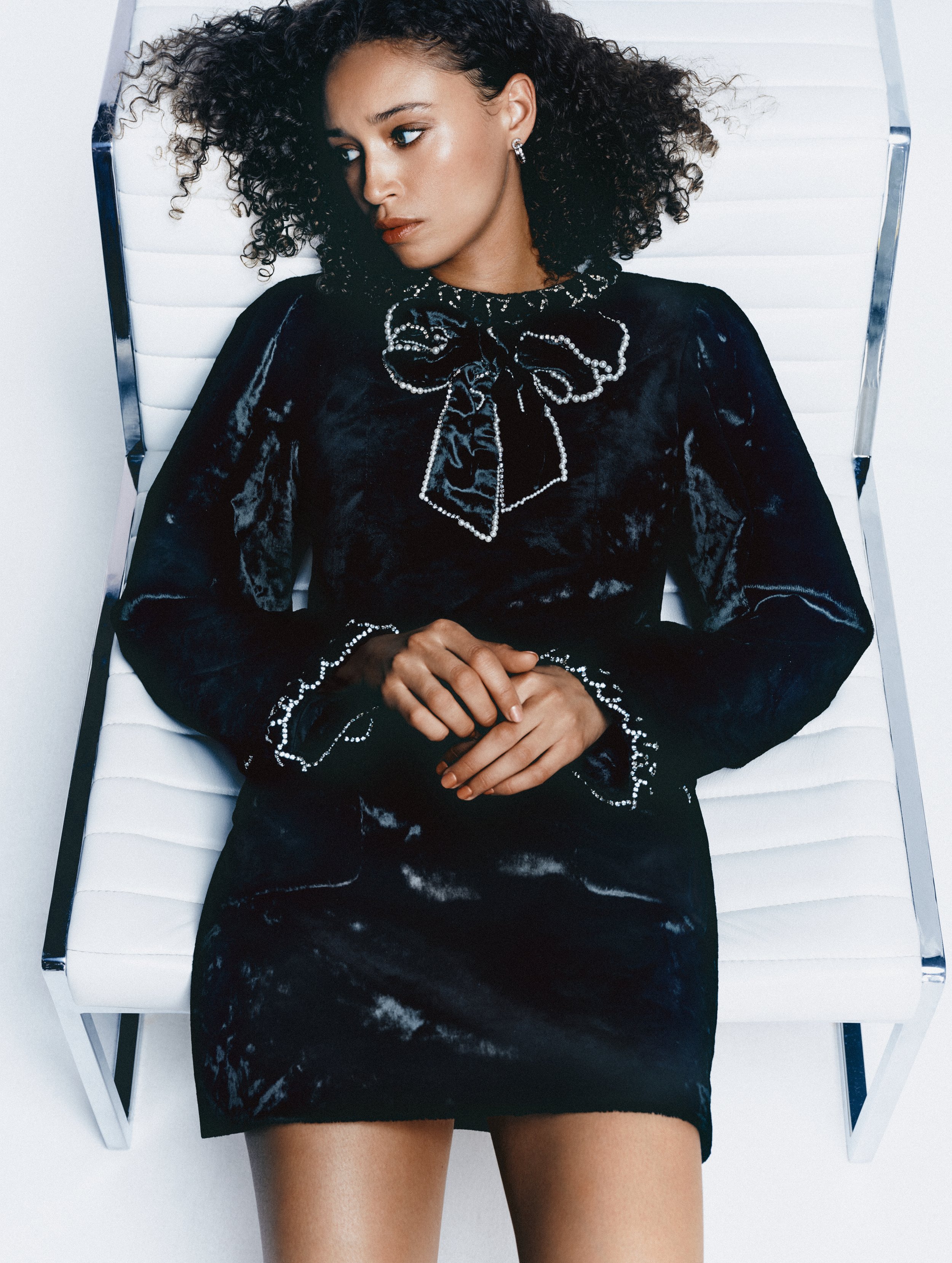 A woman lying on a white chair, wearing a black velvet dress with pearl embellishments, and has curly dark hair. She has earrings and makeup with neutral lipstick.