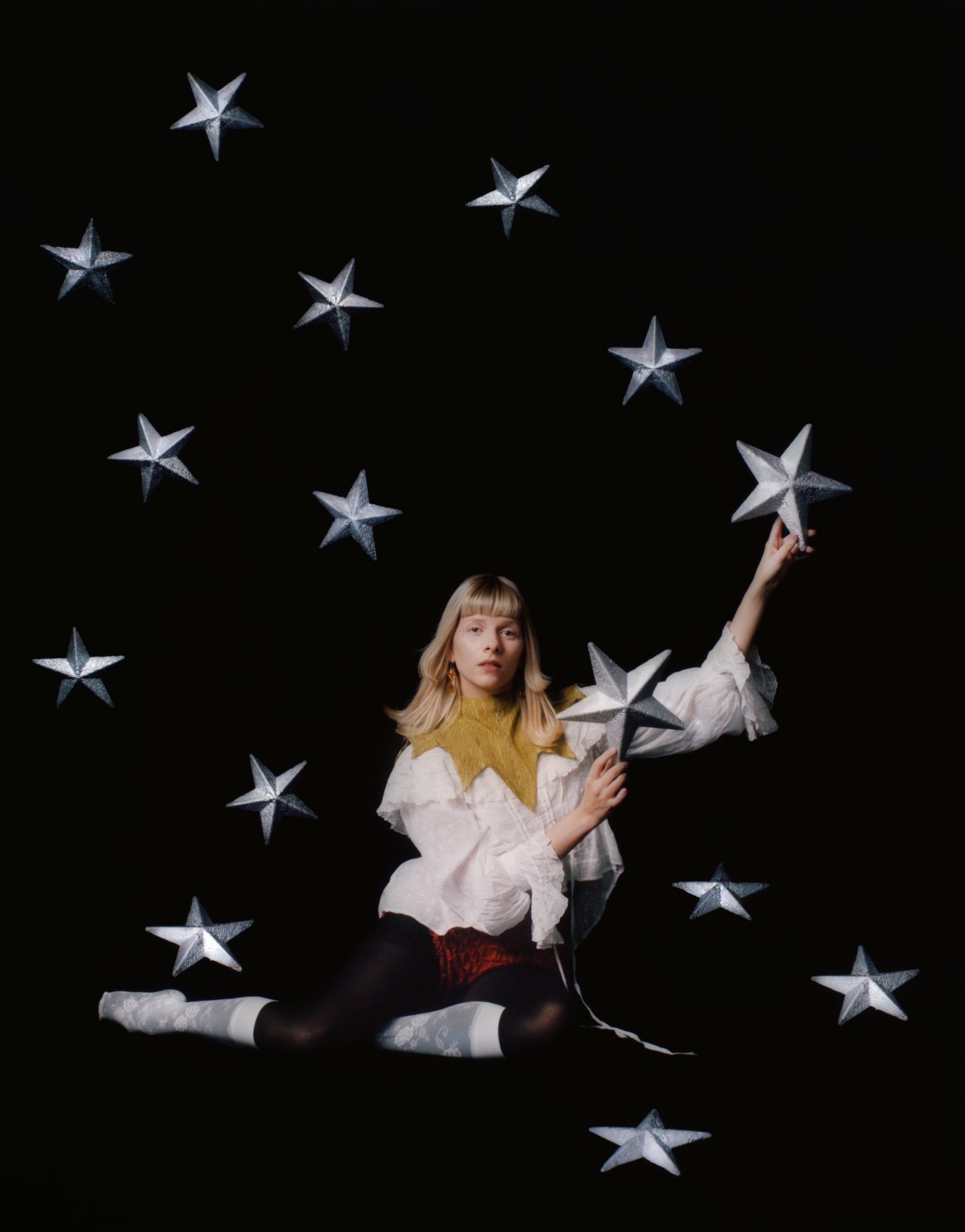 A young woman with blonde hair, wearing a yellow top and white lace stockings, poses against a black background surrounded by silver star-shaped decorations, some of which she is holding.