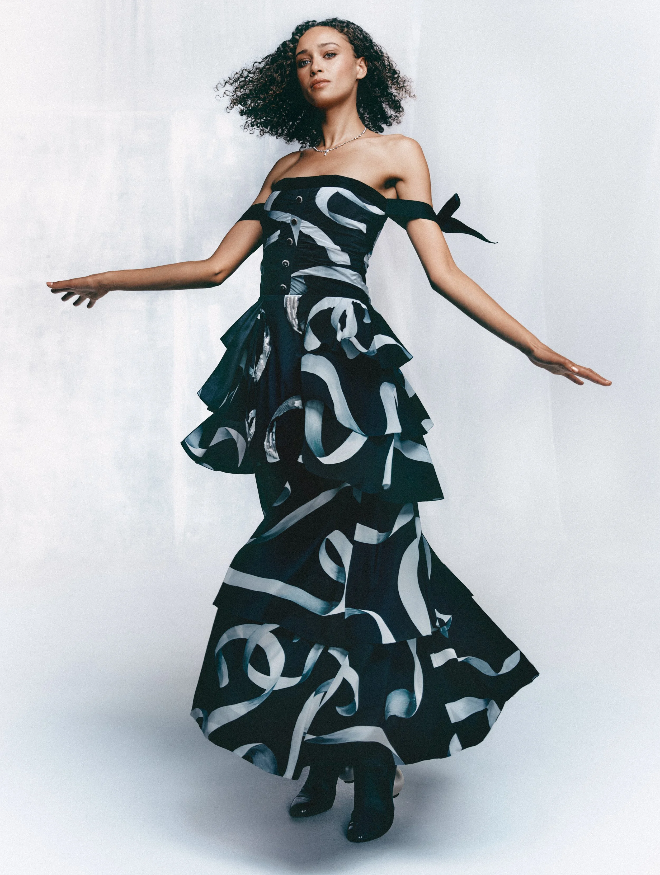 Woman with curly hair wearing a strapless black and white patterned dress, posing with arms outstretched against a light gray background.