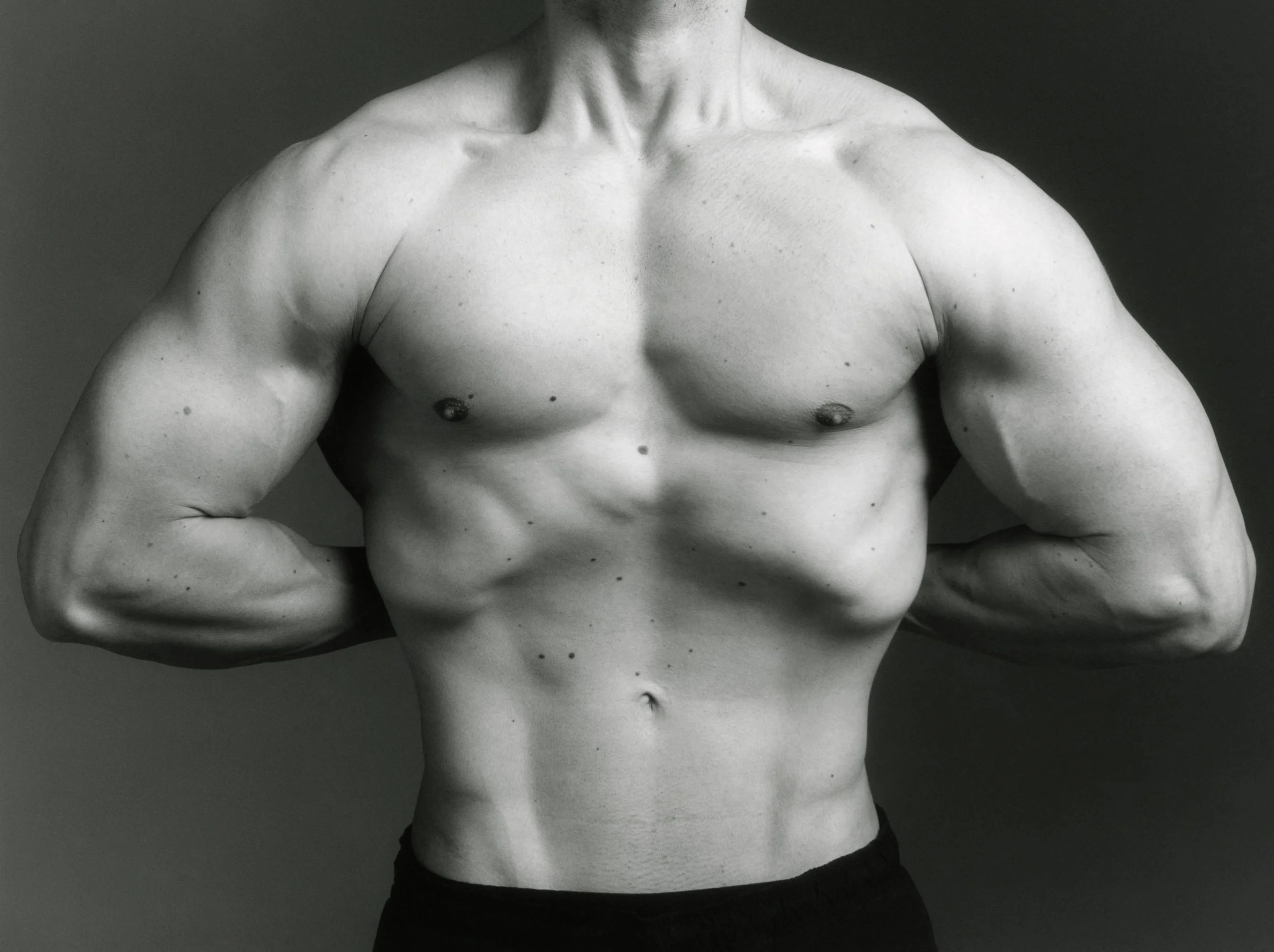 Close-up of a torso sculpture of a male with defined chest muscles, arms crossed over his abdomen, and the head not visible, set against a dark background.