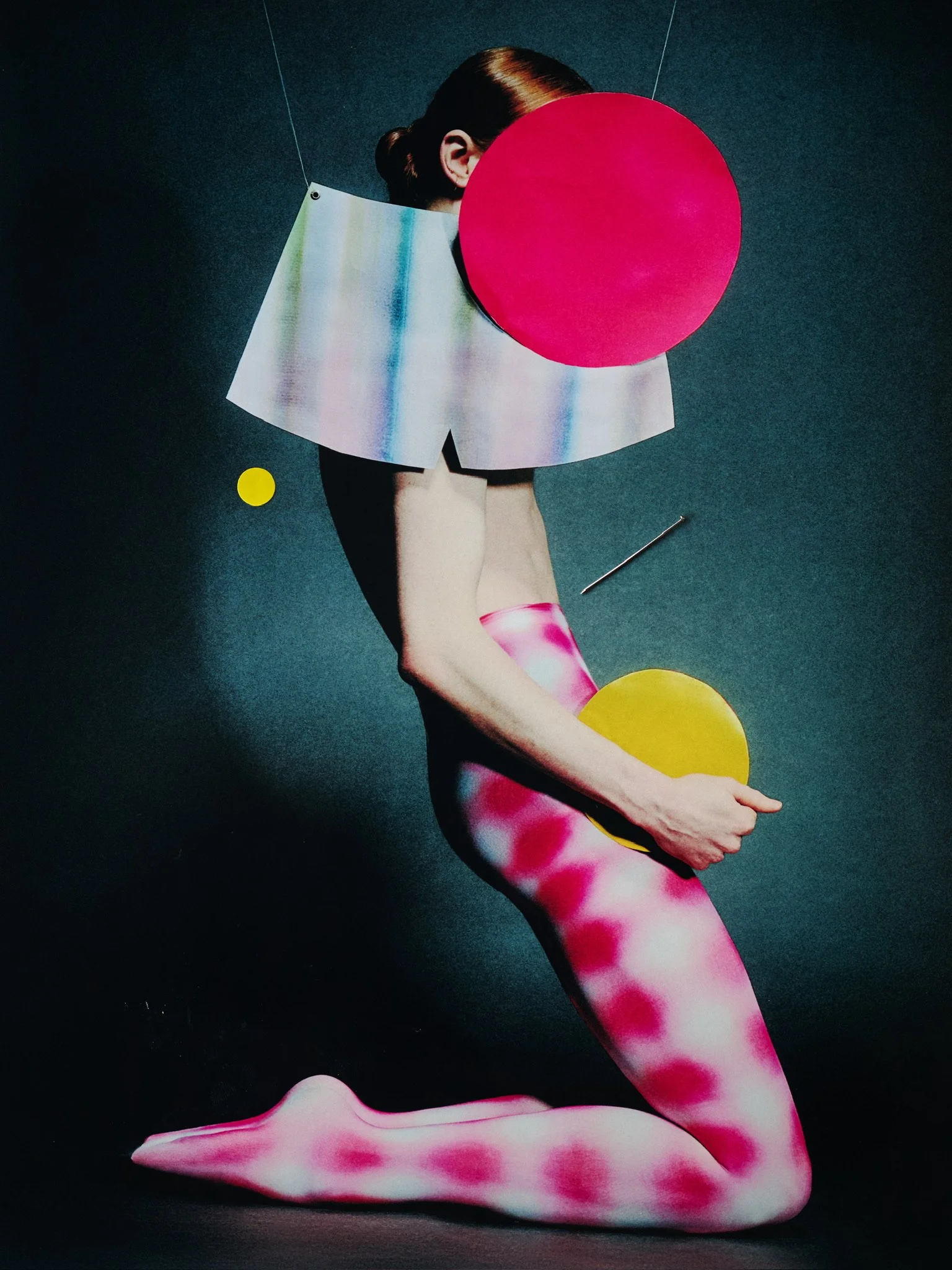A person with red hair, sitting on the floor, partially obscured by a large pink circular shape and an iridescent rectangular sheet covering the upper body. The background is dark or black.