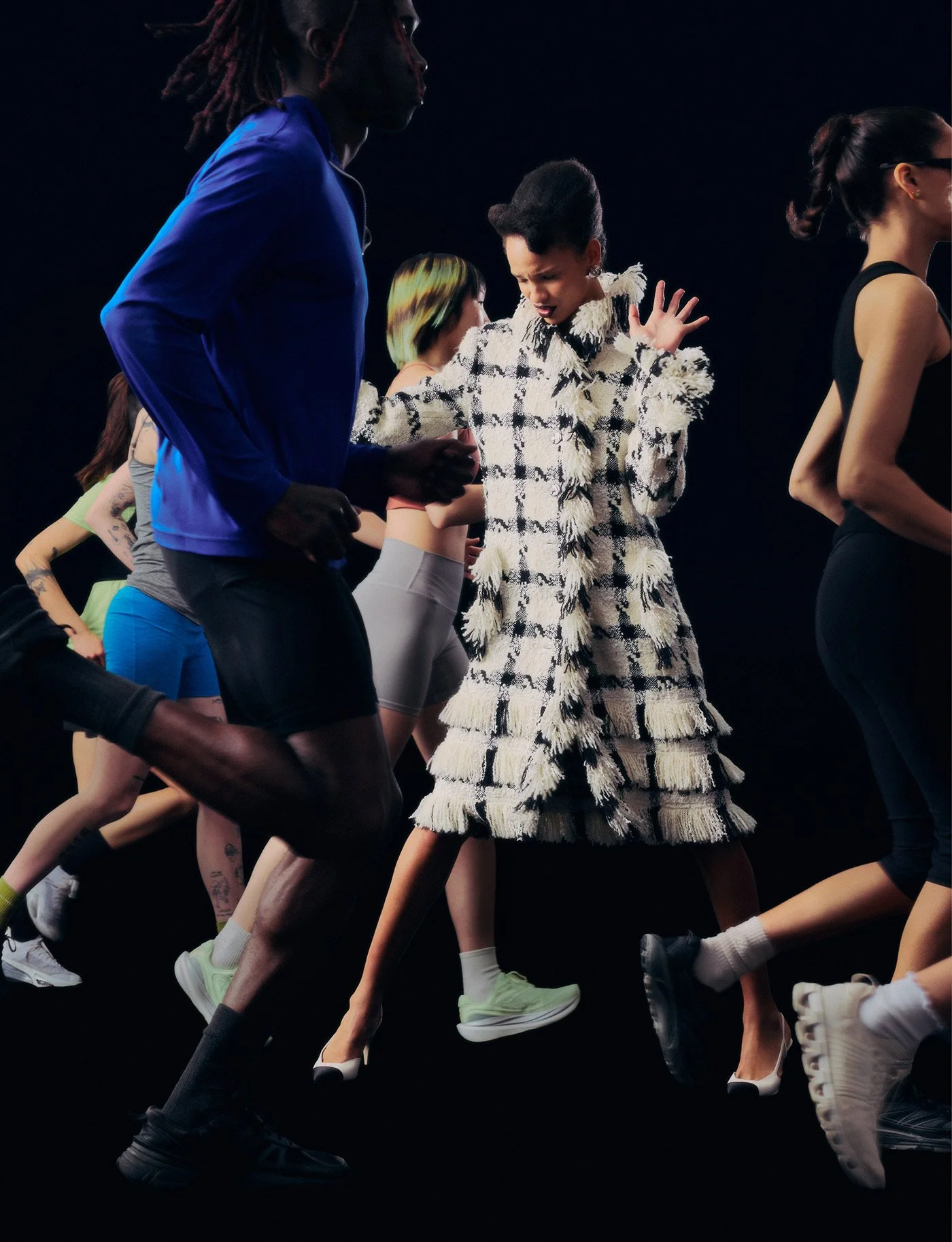 A group of diverse women running or dancing against a dark background, with one woman in the center wearing a textured, plaid coat.