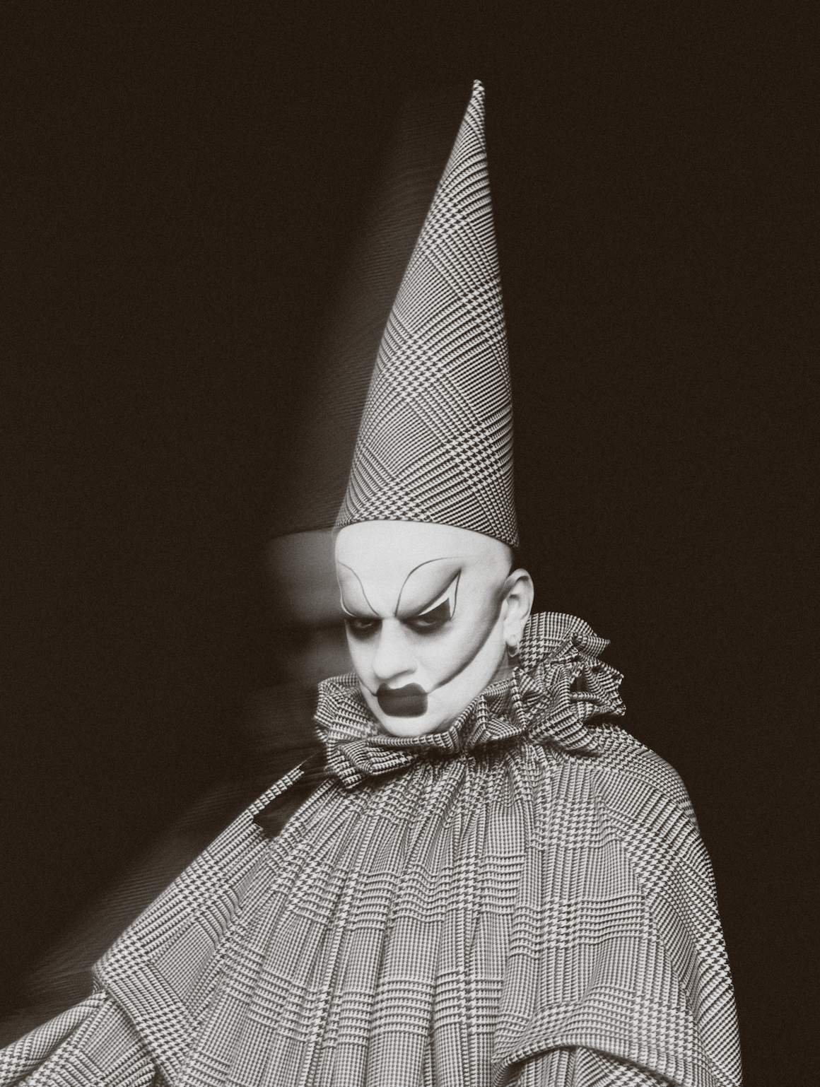A clown with makeup, wearing a pointed checkered hat and a matching checkered outfit with ruffled collar, staring downward with a serious expression.