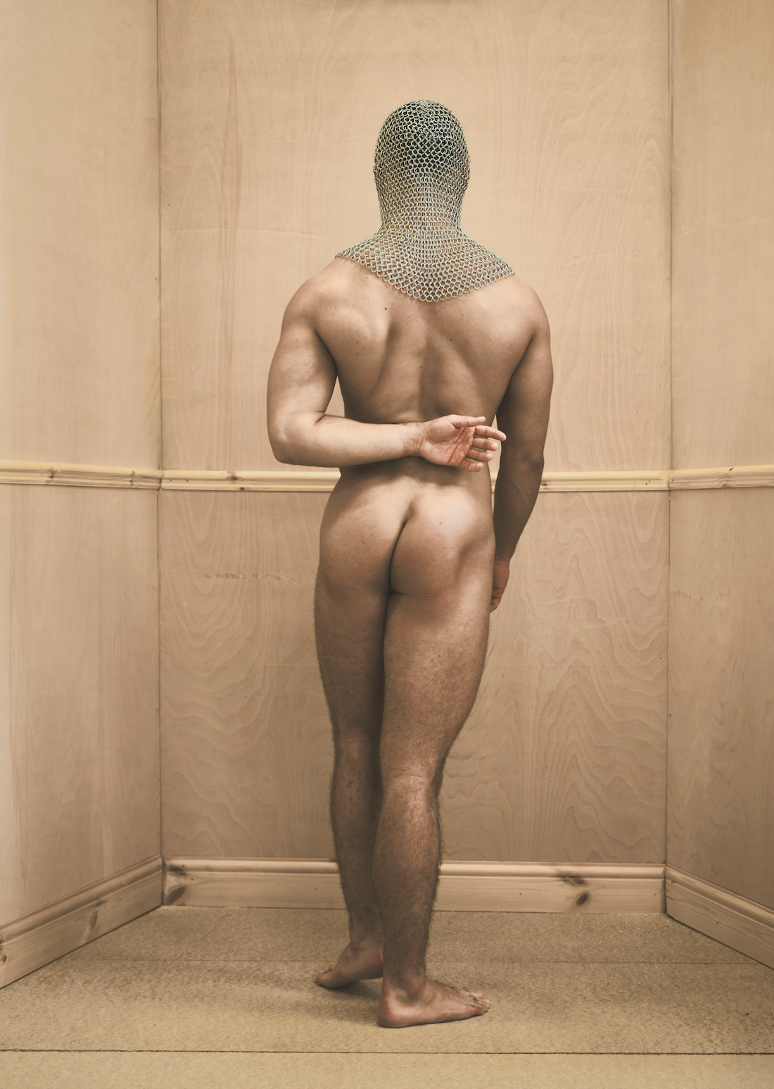 A naked man standing with his back to the camera, wearing a chainmail hood, in a wooden-paneled room.