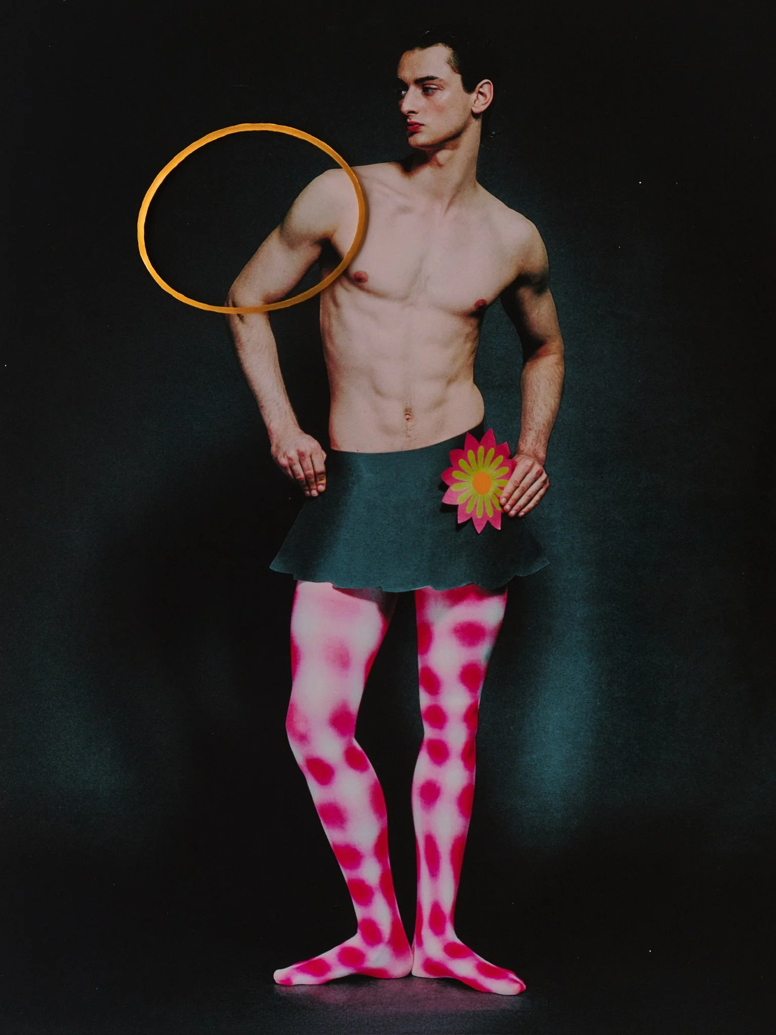 A shirtless young man with black hair standing against a dark background, wearing pink tights with red polka dots, holding a pink flower in one hand, and a yellow ring positioned around his upper arm.