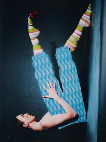 Person lying on the floor with legs raised against a wall, wearing blue striped pants and colorful striped socks, with a hand on their leg and a red object nearby.