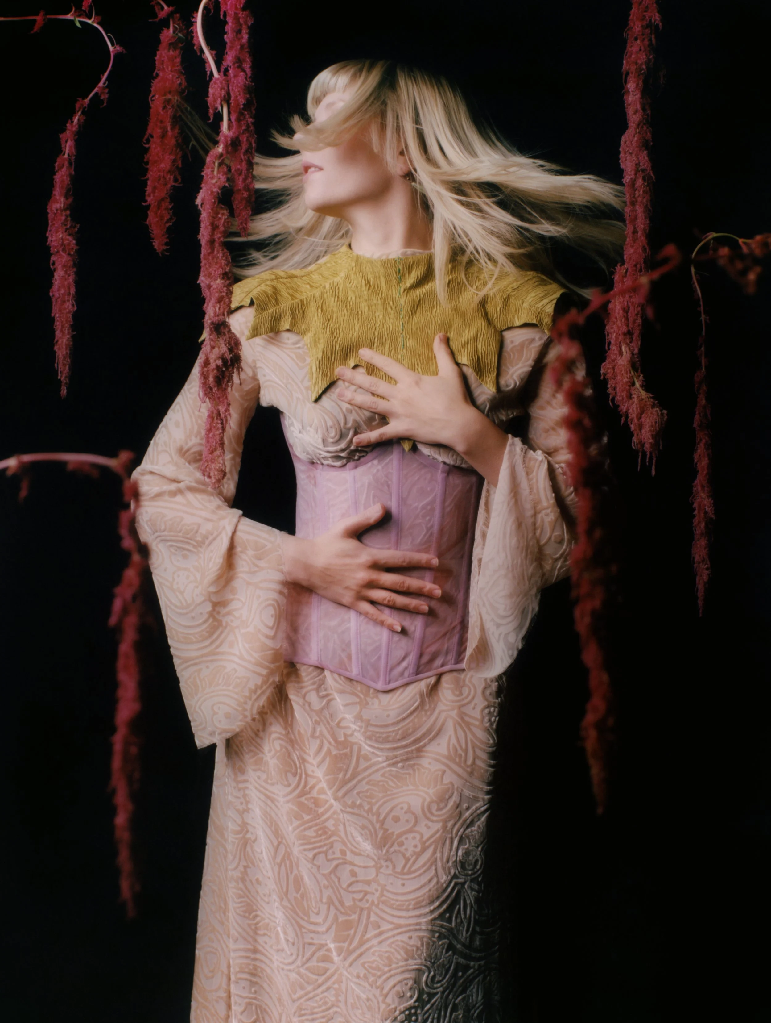 A woman with blonde hair wearing a detailed, pastel-colored gown with a yellow top and purple corset, standing with one hand on her abdomen and the other on her chest, surrounded by hanging red flowers against a black background.