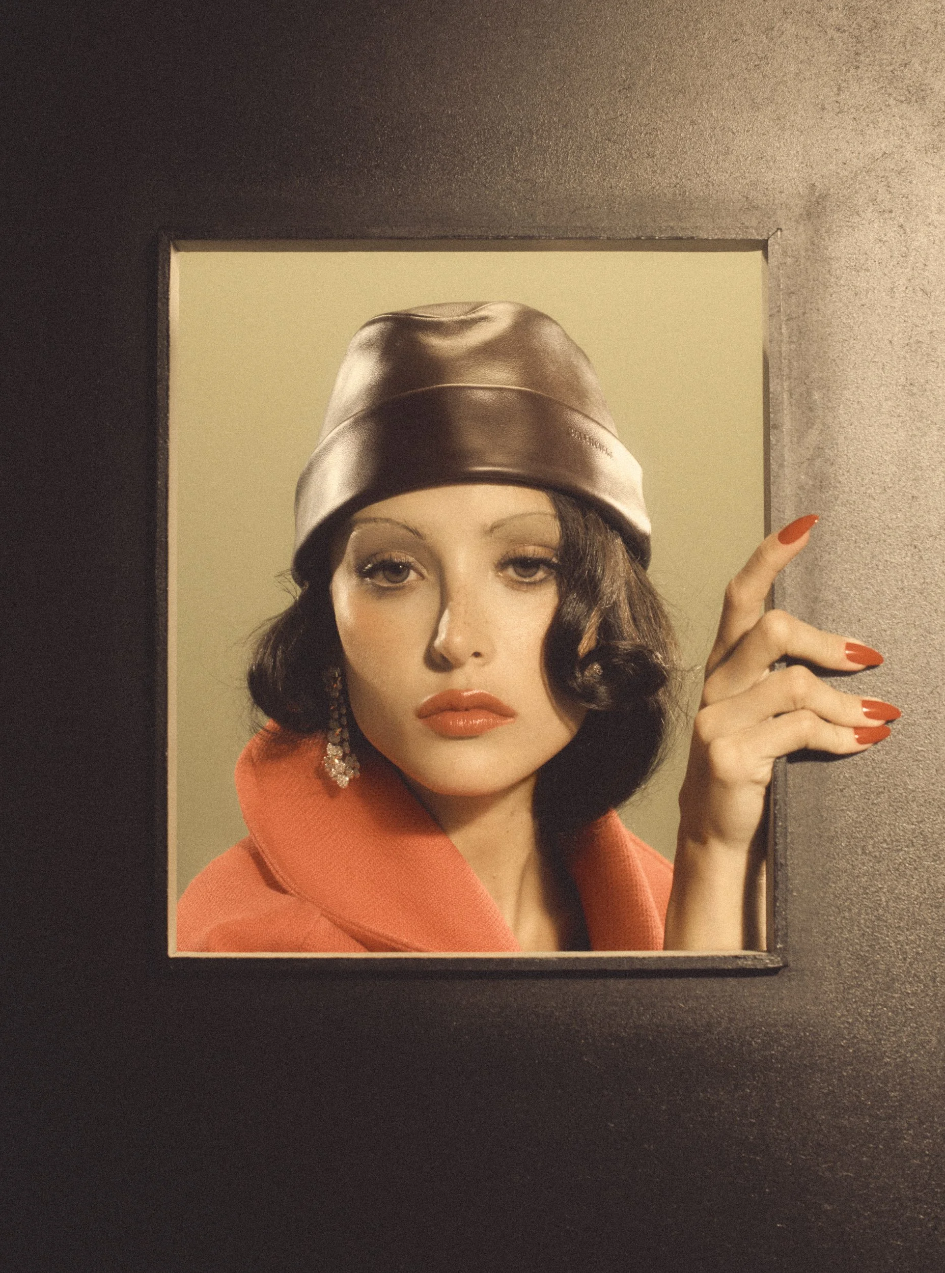 A woman with dark hair, light lipstick, wearing an orange coat, silver hat, and red nail polish, posing beside a painting or portrait of herself. She is making a gesture with her right hand, pointing upward with her index finger.