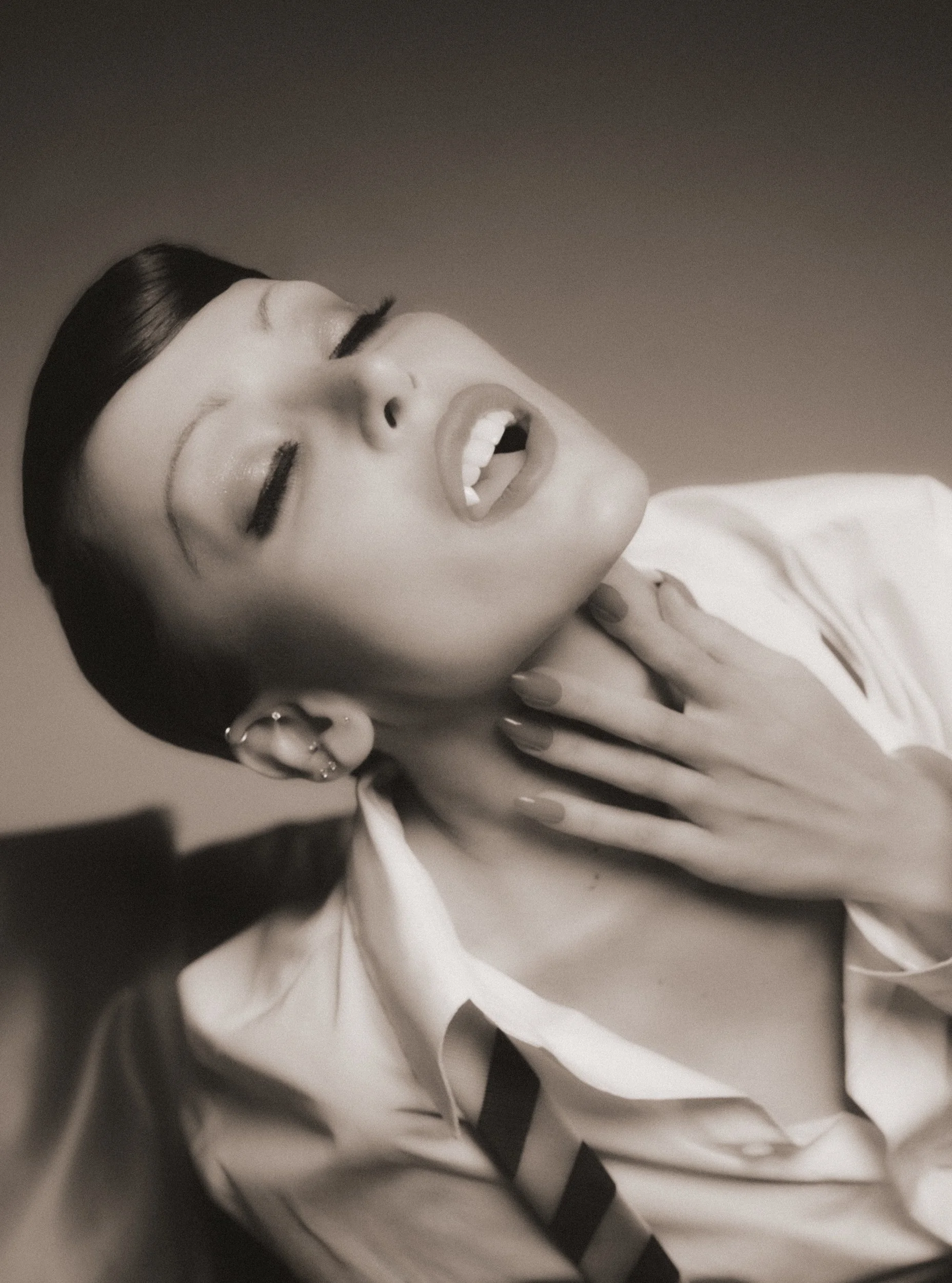A woman with dark hair, wearing earrings and makeup, poses with her eyes closed and mouth open, holding her hand to her neck, dressed in a light-colored shirt with black accents.
