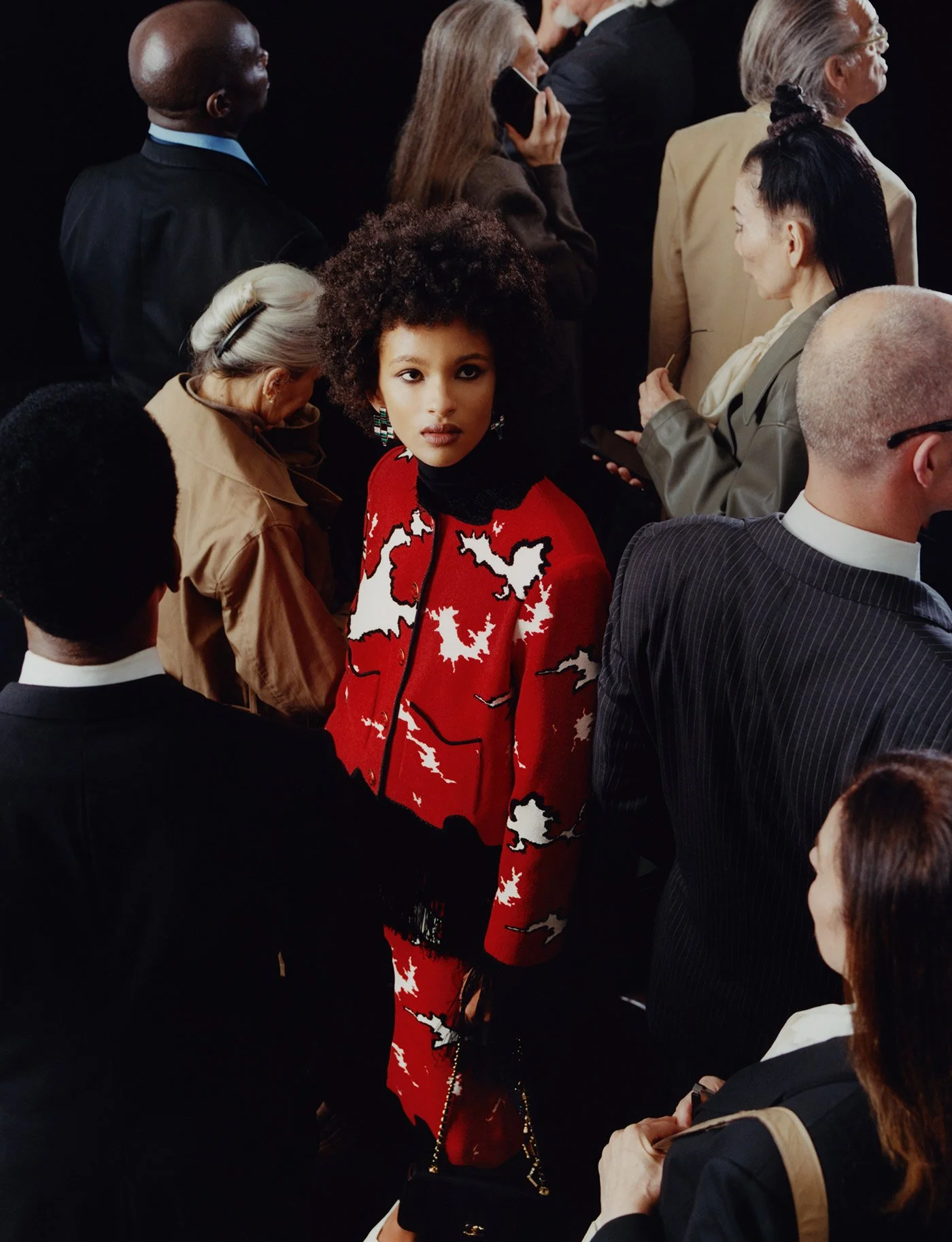 A diverse group of people, including a woman with curly hair wearing a red and white patterned outfit, are gathered together, some engaging with their phones, in a social setting.