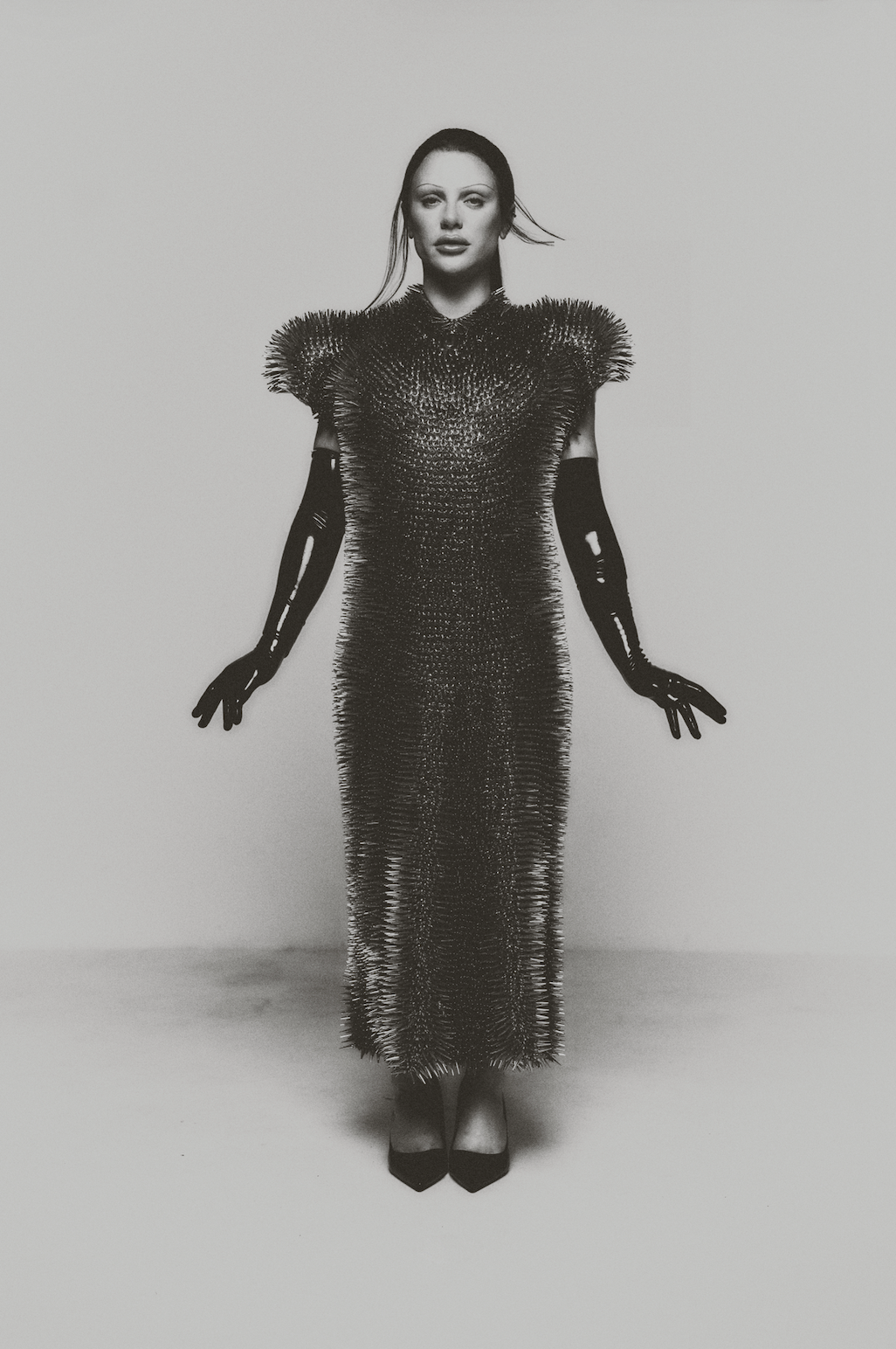A woman in a form-fitting, textured dress with shoulder pads, wearing long gloves, standing against a plain background.