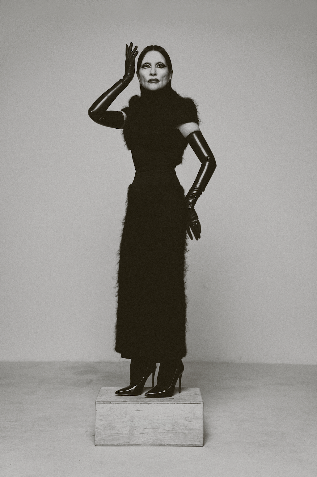 A woman with long hair standing on a small wooden block, wearing a long black dress with fur and shiny black gloves and high heels, posing against a plain background.