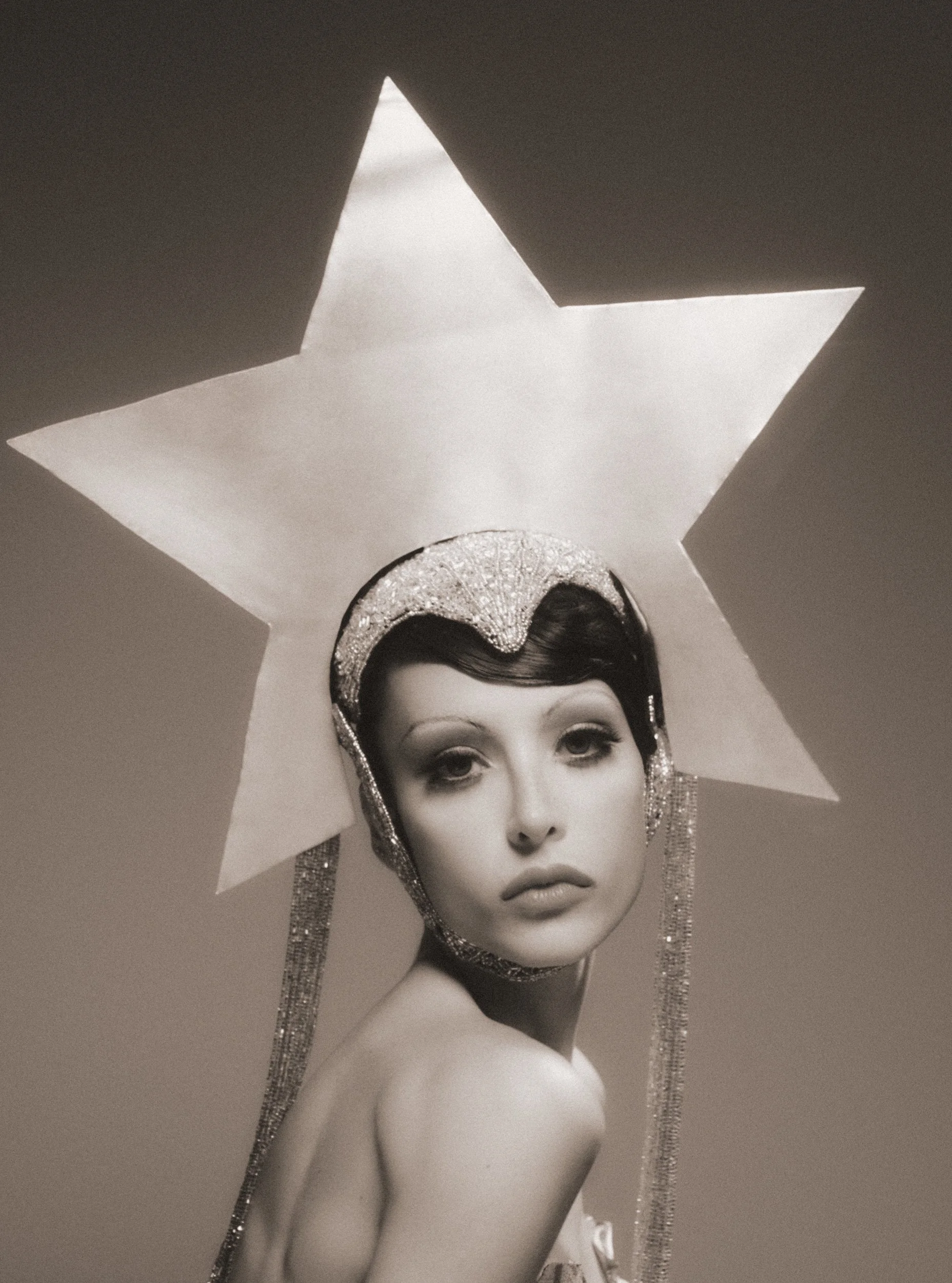 Black-and-white photo of a mannequin with styled hair, wearing a shiny headpiece and earrings, partially covered by a large star-shaped cutout in the ceiling.