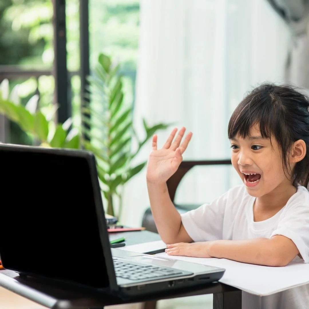 A young girl with dark hair in a ponytail waving and smiling at a laptop in a bright room with green plants in the background.