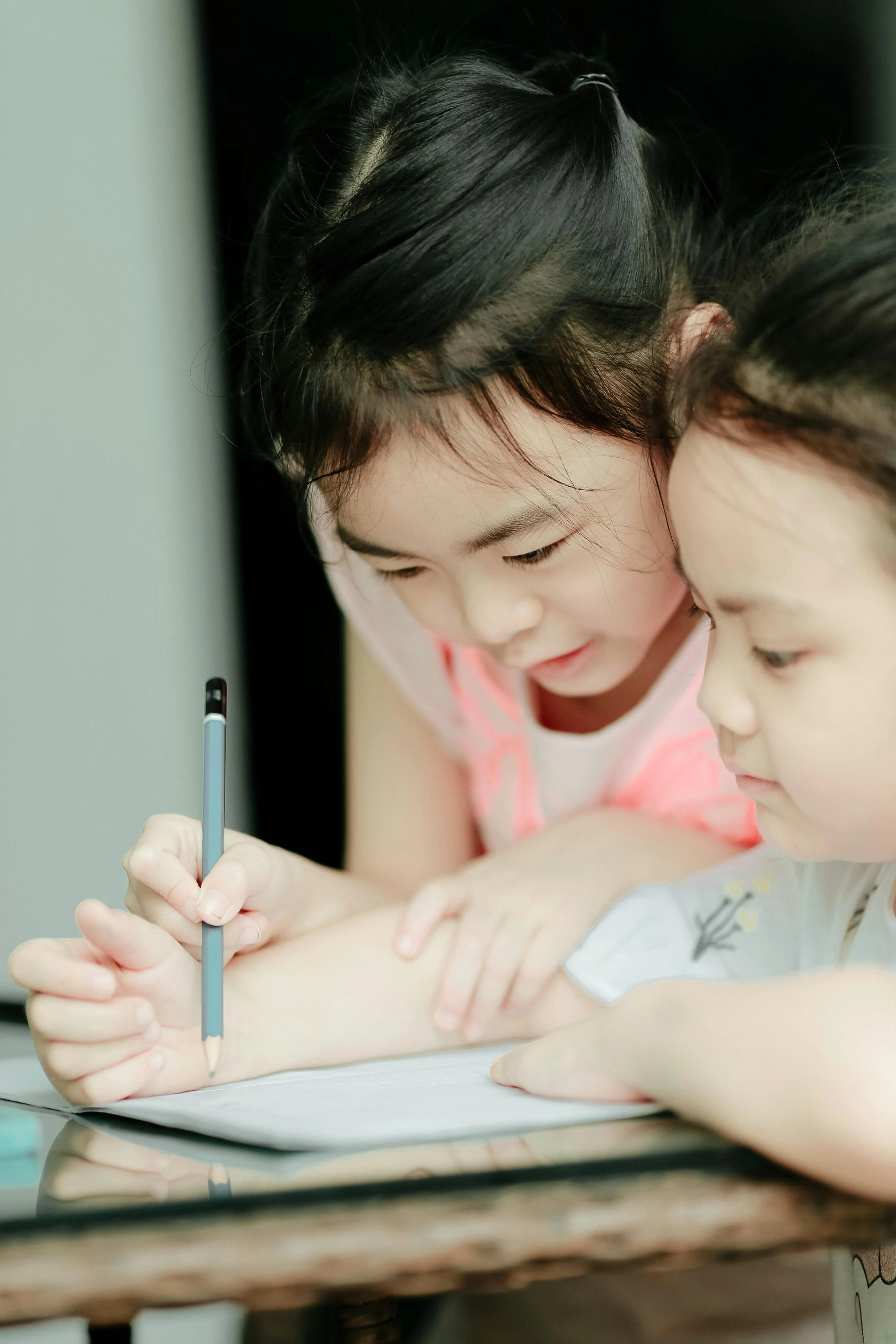 Two young girls with dark hair leaning close together as one writes in a notebook with a light-colored pen.