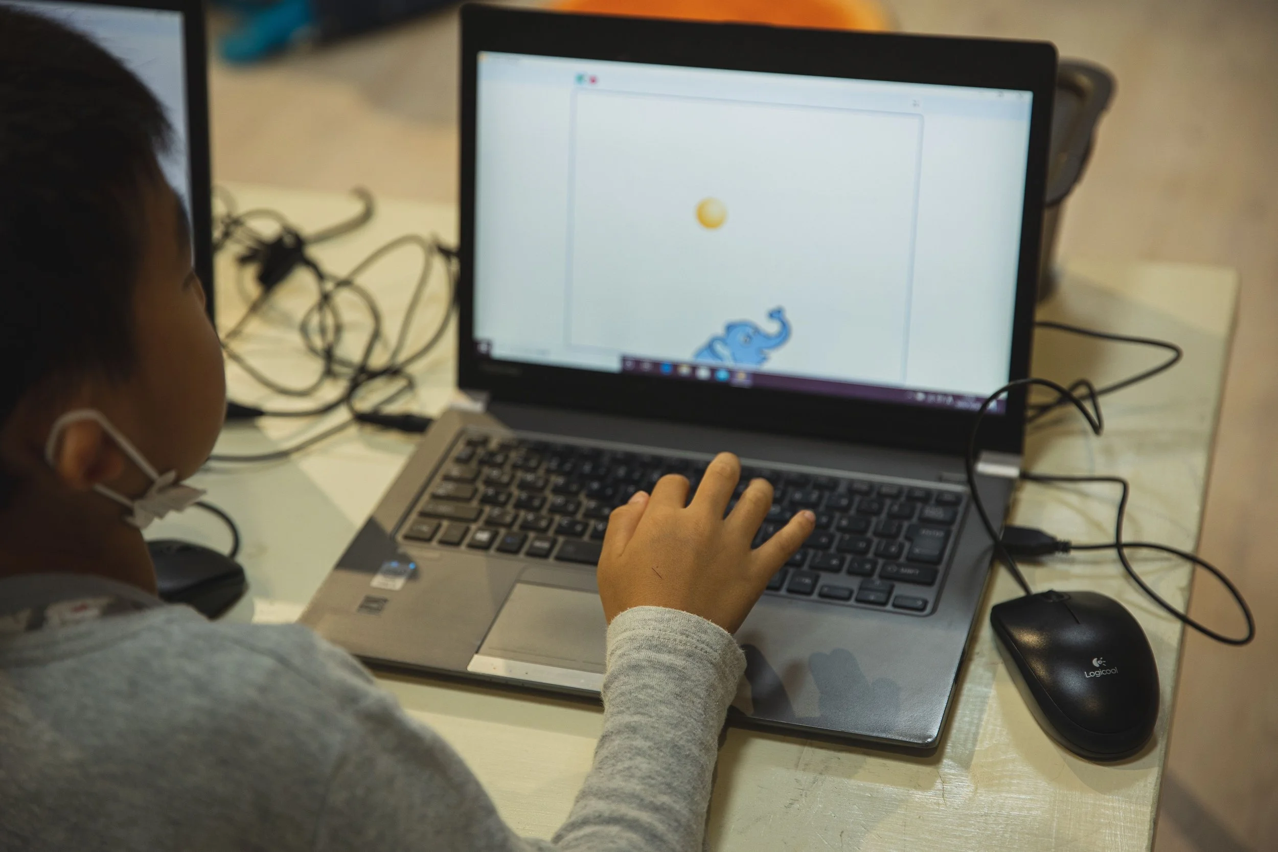 A child wearing a gray long sleeve shirt and a face mask working on a laptop. The laptop screen displays a drawing of a blue animal, possibly a dinosaur, with a yellow ball above it. There are various tangled cables and objects on the table.