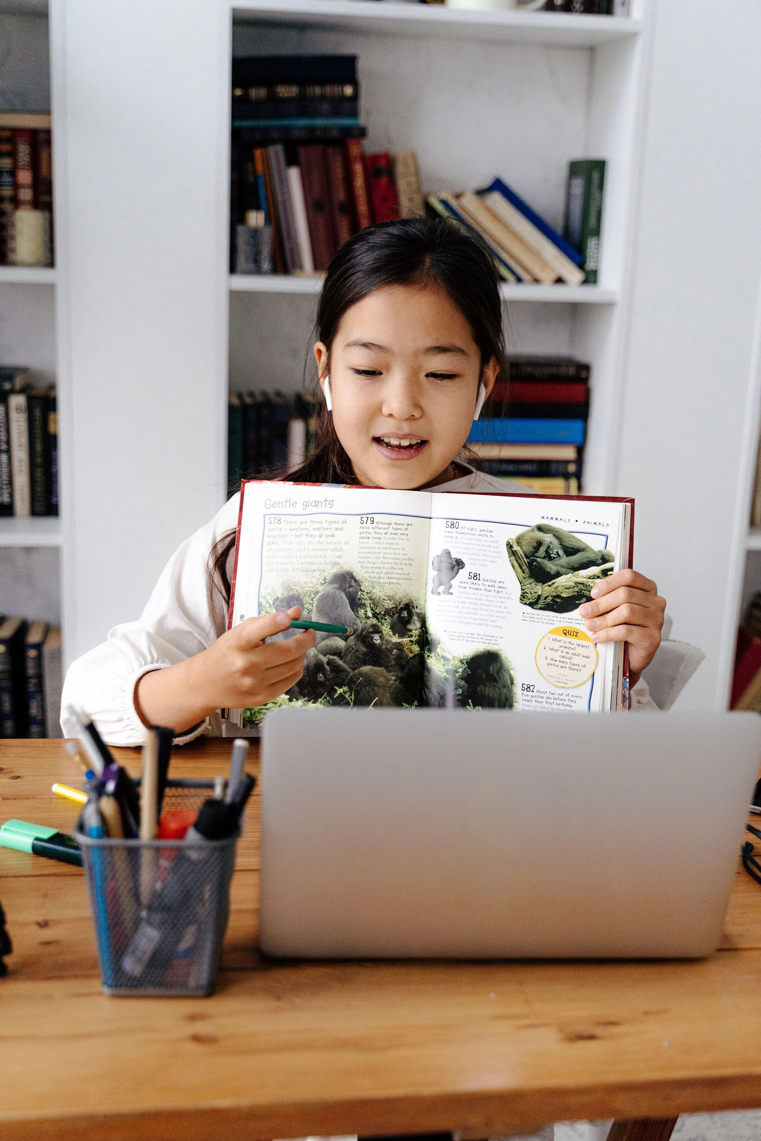 A young girl with dark hair and light skin sitting at a wooden desk in front of a laptop. She is reading a colorful book about animals, with a pen in her hand. Behind her, there is a white bookshelf filled with books, and a container with colored markers on the desk.