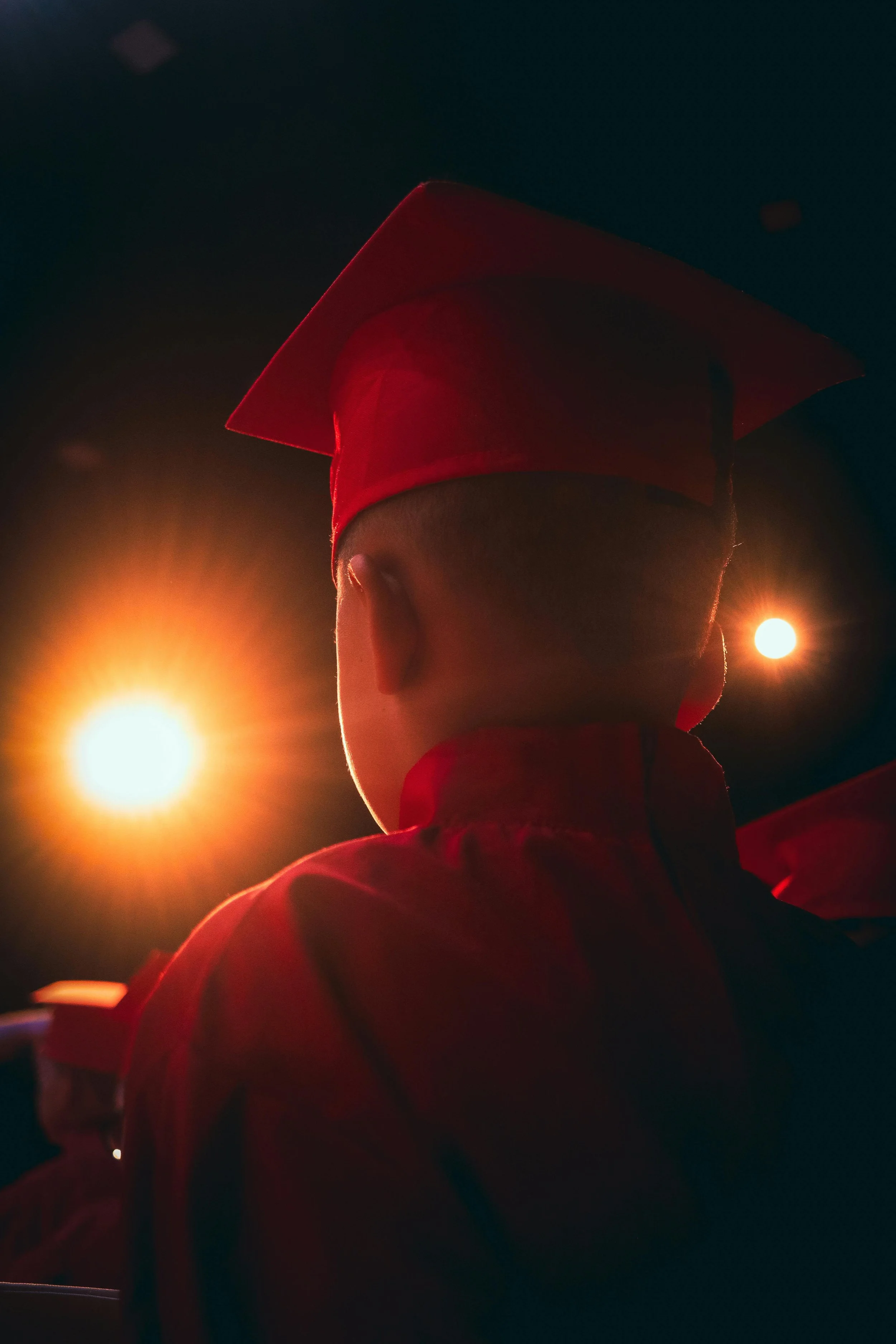 Person in a graduation cap and gown with back turned, illuminated by bright orange and yellow lighting, against a dark background.